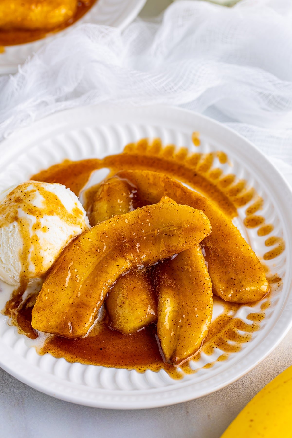 Overhead upclose angle of New Orleans Bananas Foster with scoop of vanilla ice cream, on a white plate. White gauze linen in the background and banana.