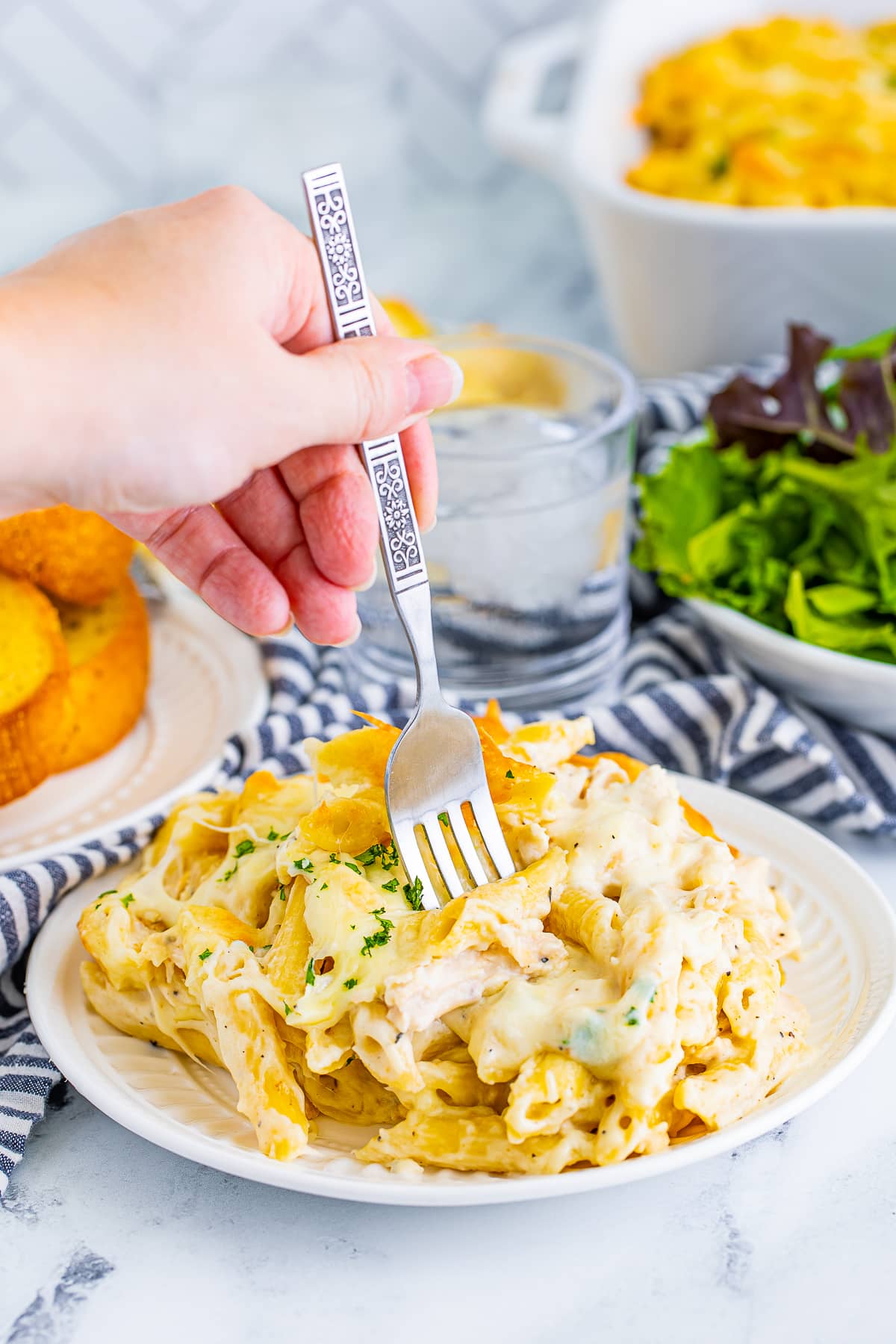 A silver patterned fork in hand grabbing some of Chicken Alfredo Bake on a plate with glass of water, bowl of salad, and navy striped linen in the background.