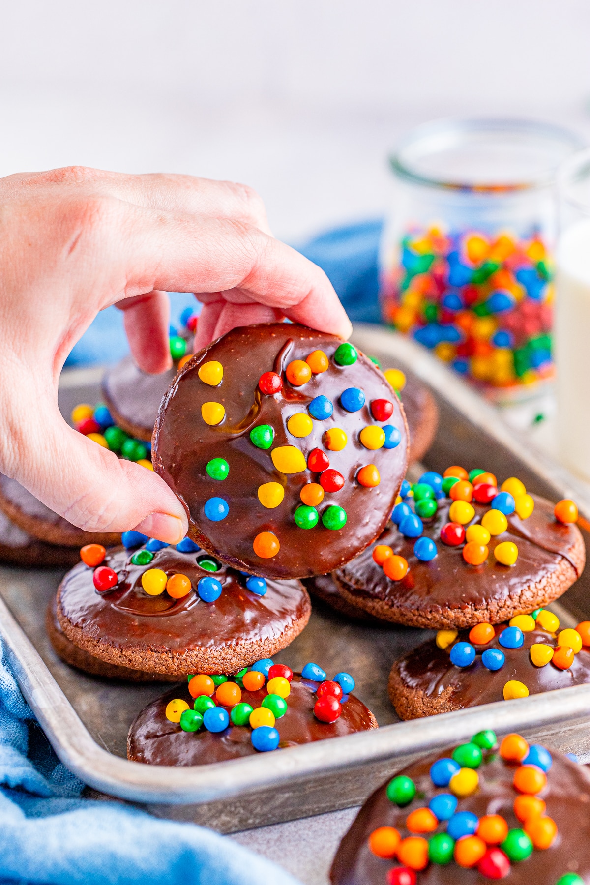 A Cosmic Brownie Cookie being held up by a hand to see the topping. Glass of sprinkles, glass of milk, and blue linen in the background. More cookies are sitting on a silver metal tray.