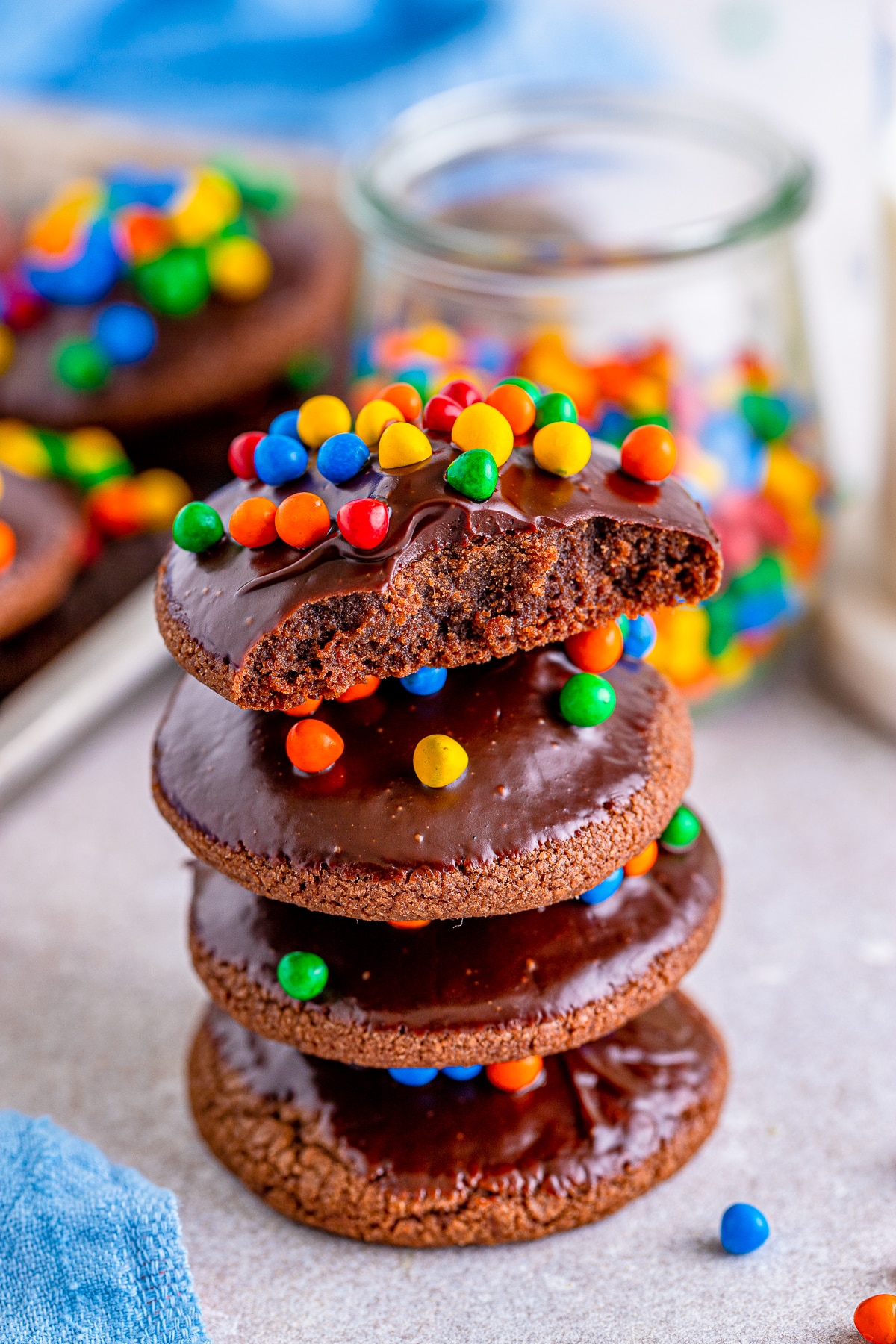 A stack of Cosmic Brownie Cookies on a concrete backdrop, glass of sprinkles in the background with blue linen