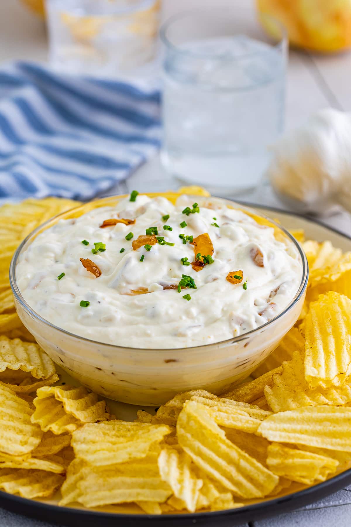 French Onion Dip Recipe in a clear glass bowl surrounded by wavy potato chips with glass of water and blue striped linen straight on angle.