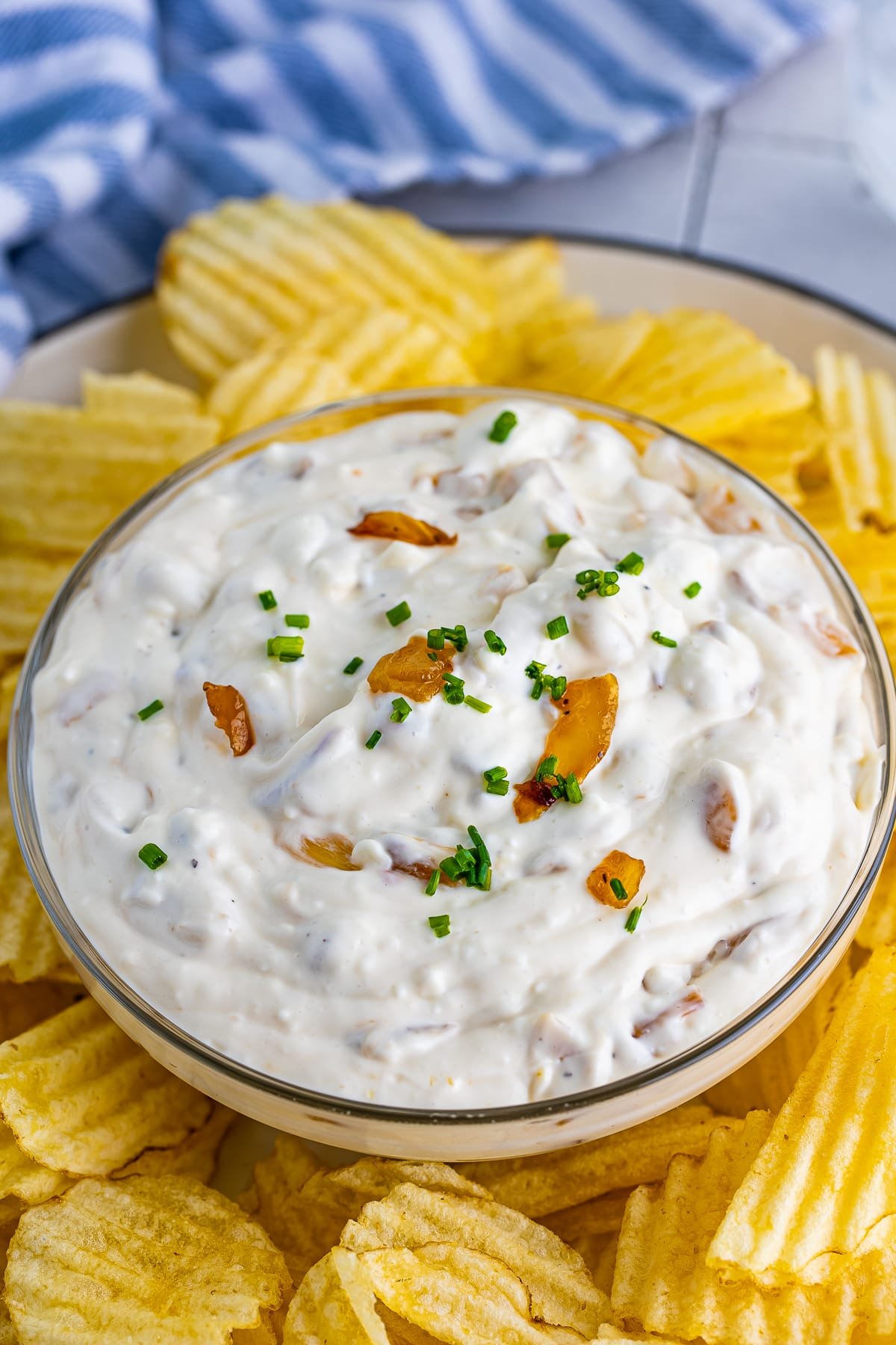 Angled shot of French onion dip recipe in a clear glass bowl surrounded by wavy potato chips, and blue striped kitchen towel