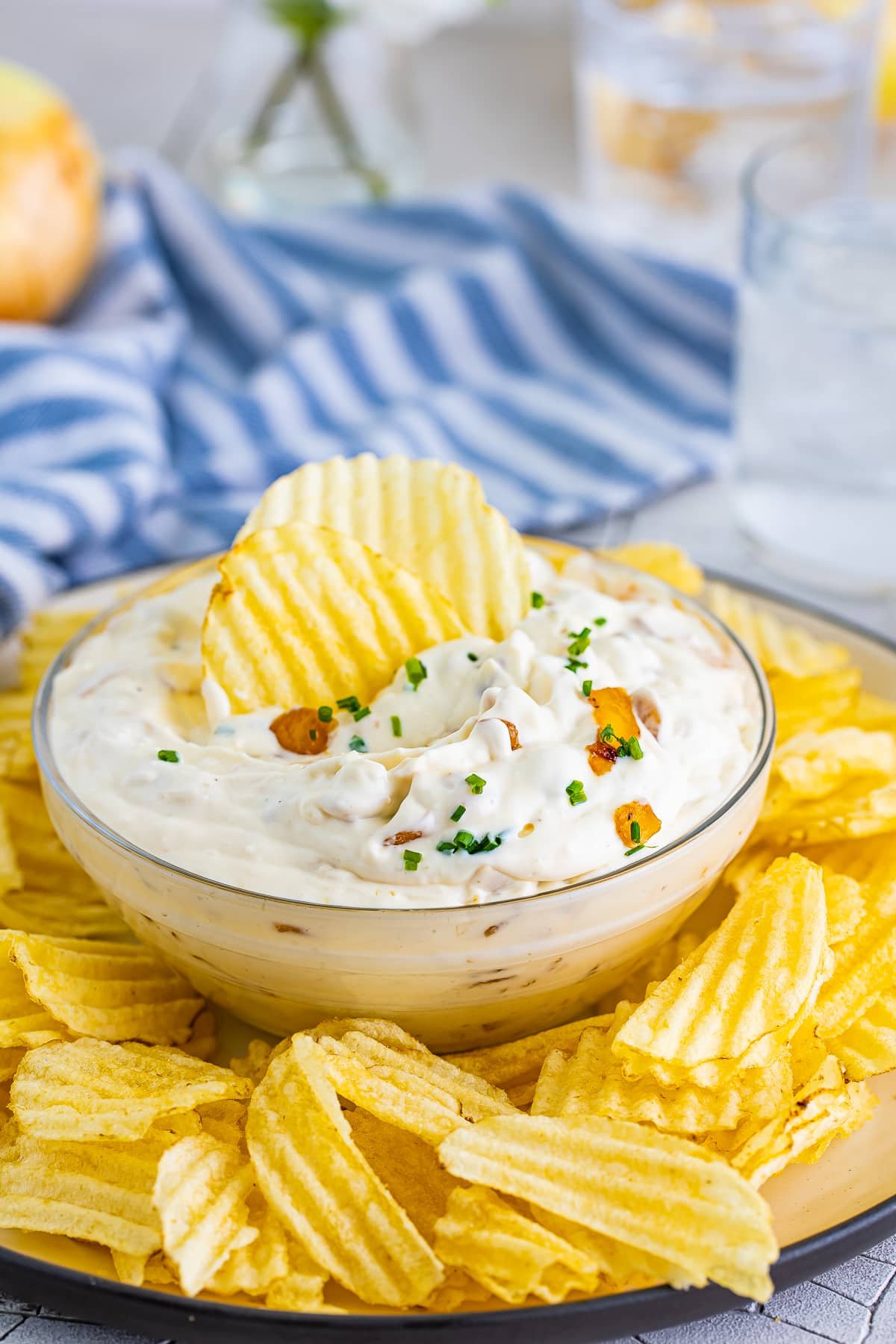 French onion dip recipe in a clear glass bowl with two wavy potato chips sticking out of it. Blue striped linen and glasses of ice water in the background