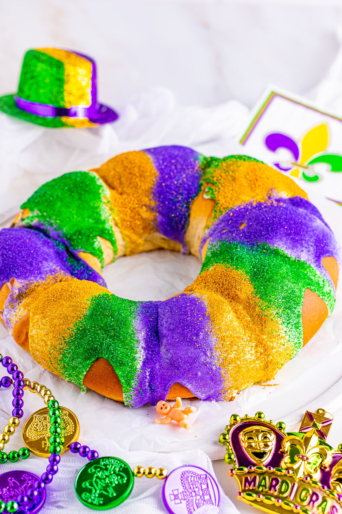 Full shot of King Cake Recipe served on a white serving platter with Mardi Gras decorations and white backdrop.