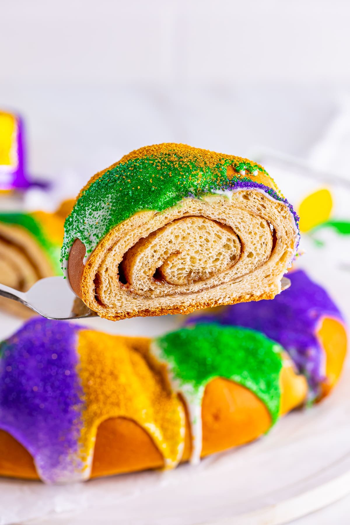 A slice of this King Cake Recipe on a cake server over the whole cake with white background.