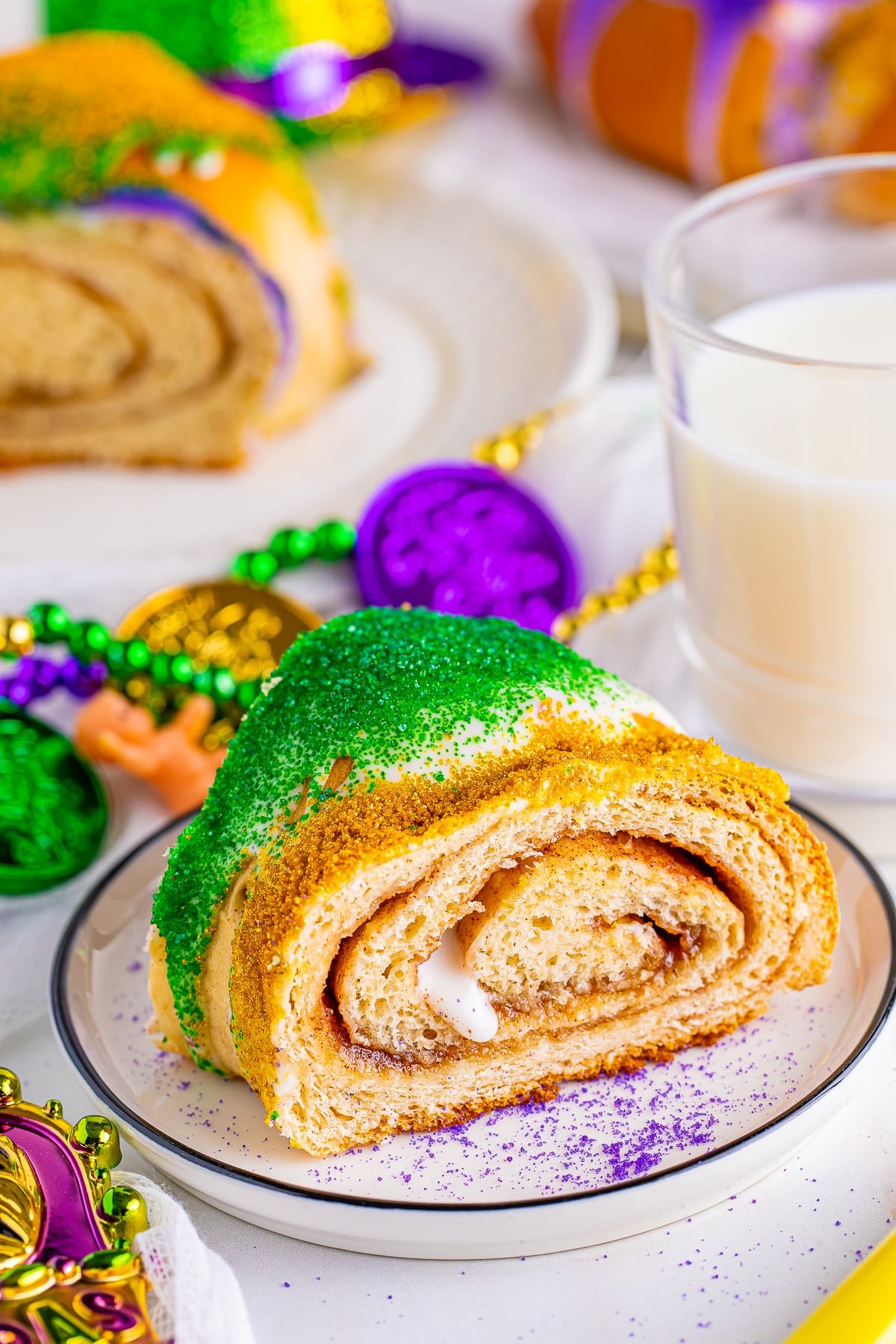 A slice of this King Cake Recipe on a white plate with black rim, mardi gras decorations in the background with a glass of milk.