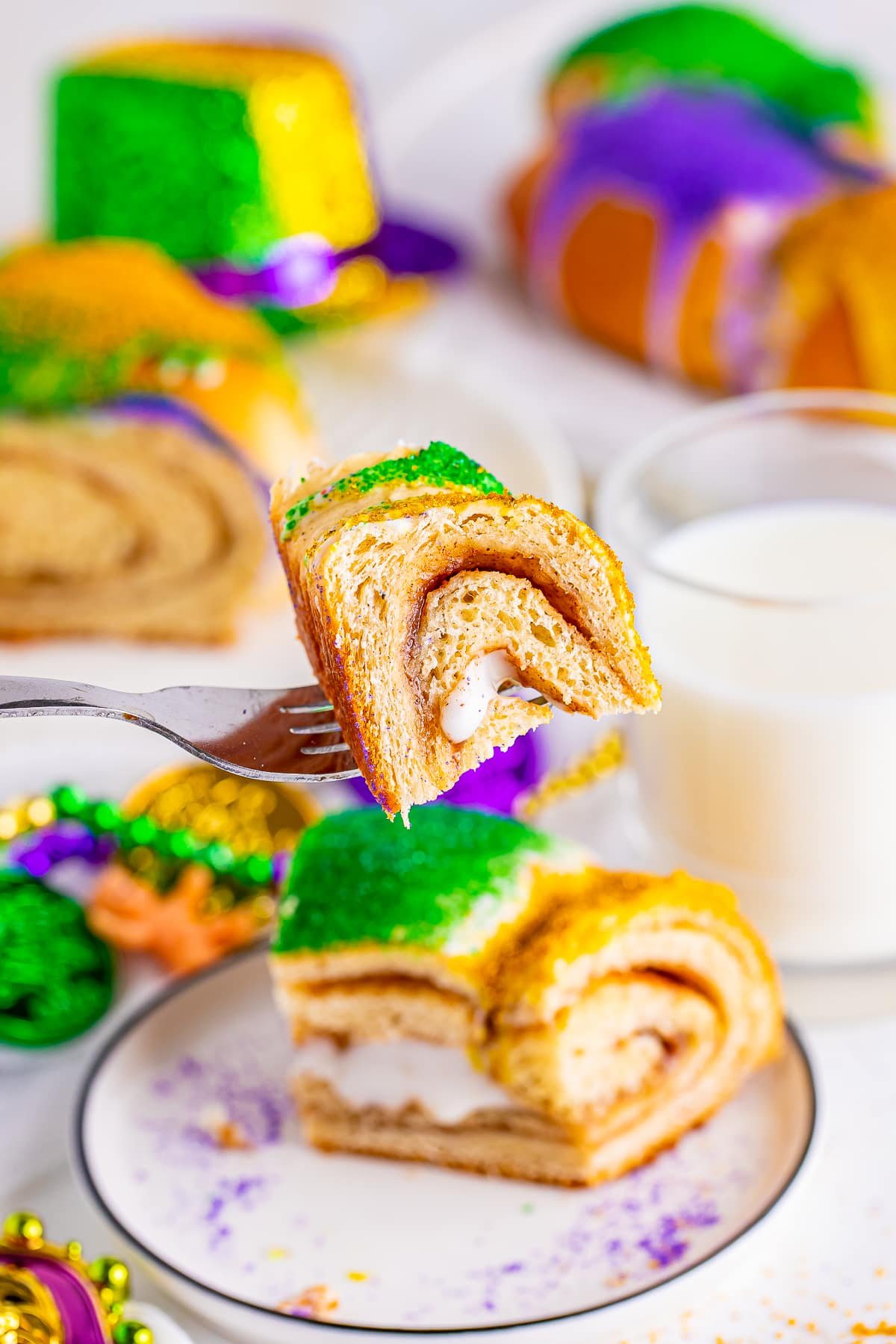 A bite of this King Cake Recipe on a fork suspended in air, you can see slices of the cake, a glass of milk, and mardi gras decorations in the background.