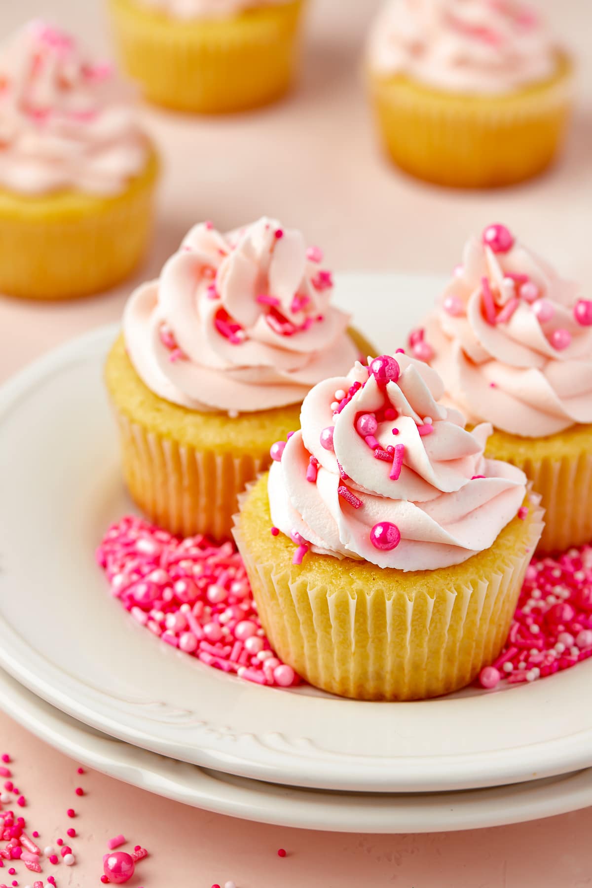 Three Valentine's Day Cupcakes sitting on a white plate with pink sprinkles scattered around on a pink tabletop. Pink frosting and pink sprinkles on the cupcakes
