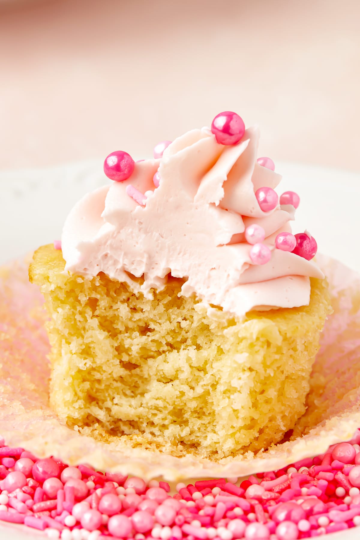 Photo showing an interior shot of Valentine's Day Cupcakes with a bite taken out. Pink frosting and pink sprinkles on the cupcake. Sittin on a white plate with more pink sprinkles surrounding the cupcake.