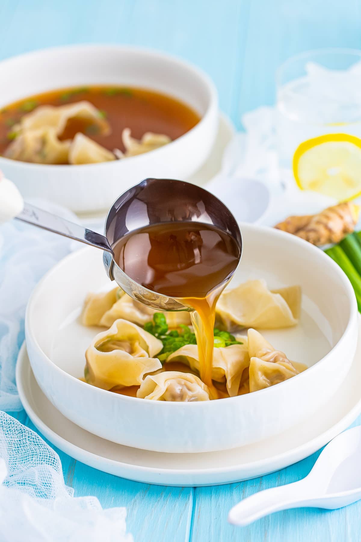 Wonton Soup Recipe showing the broth being poured into a white bowl over wontons with a bright blue backdrop.
