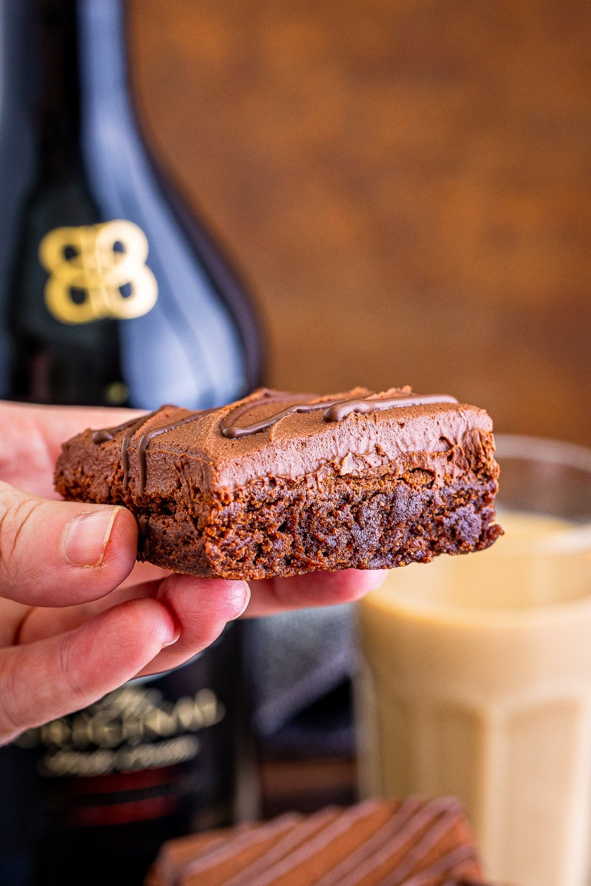 A hand holding a Bailey's Brownie in mid air, bottle of alcohol in the background with brown backdrop