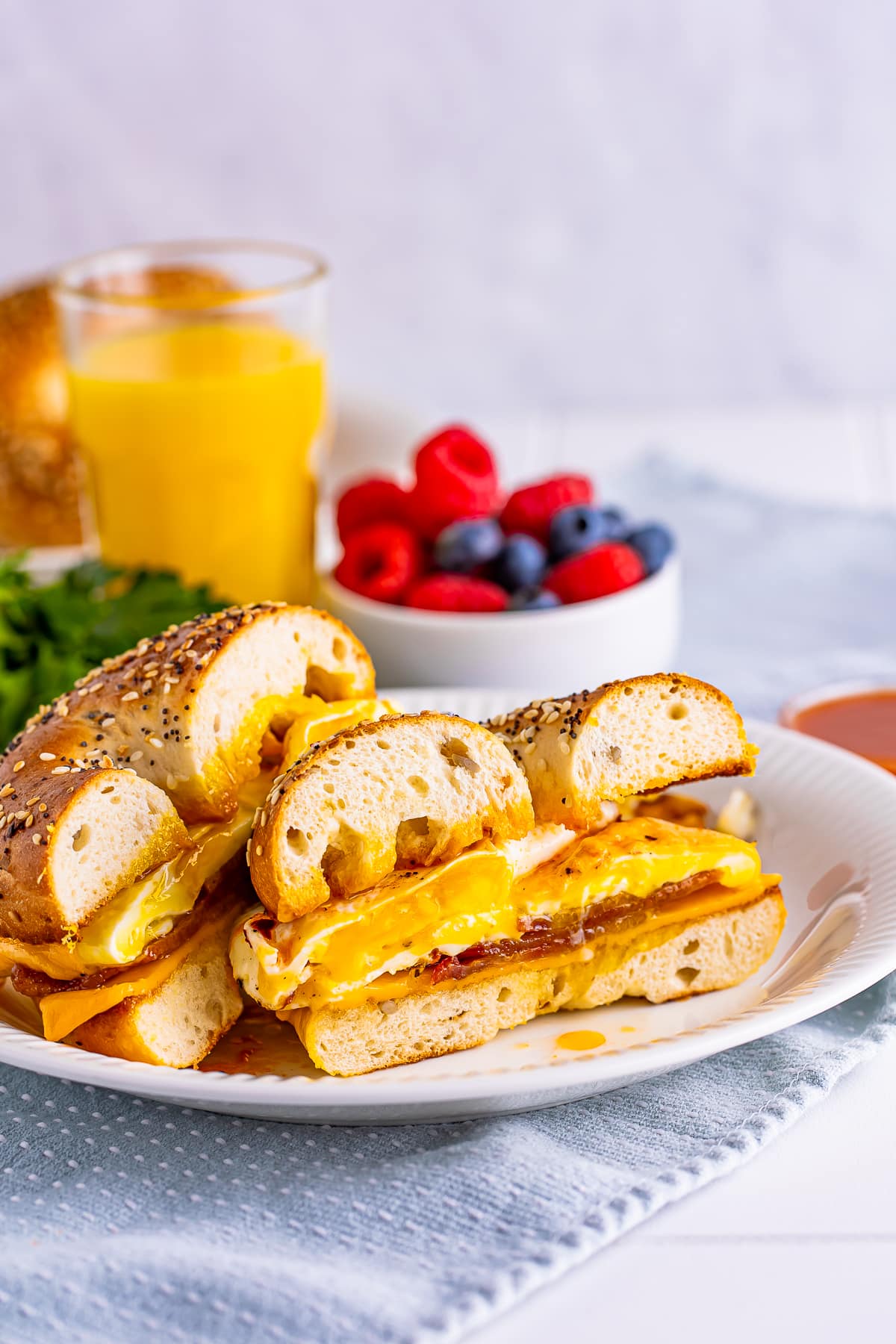 Interior slice shot of Breakfast Bagel Sandwich with drippy egg yolk on white plate, light blue linen, glass of orange juice, small bowl of berries
