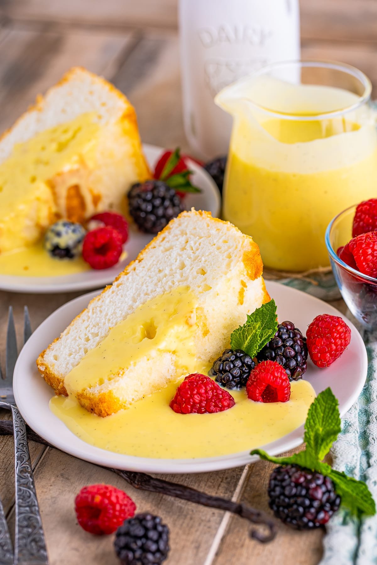 A slice of angel food cake on a white plate with berries and mint, Creme Anglaise Recipe is pooled on the plate and drizzled over the cake. Served on a wooden table top with mint and milk jug in the background.