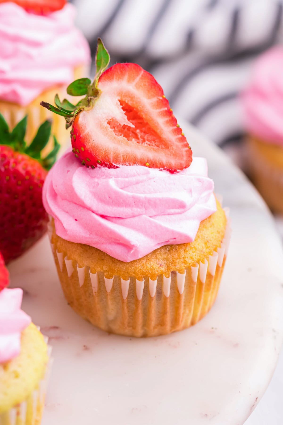 45 degree angle showing a Strawberry Cupcakes sitting on a white marble cake stand with other cupcakes in the back ground.