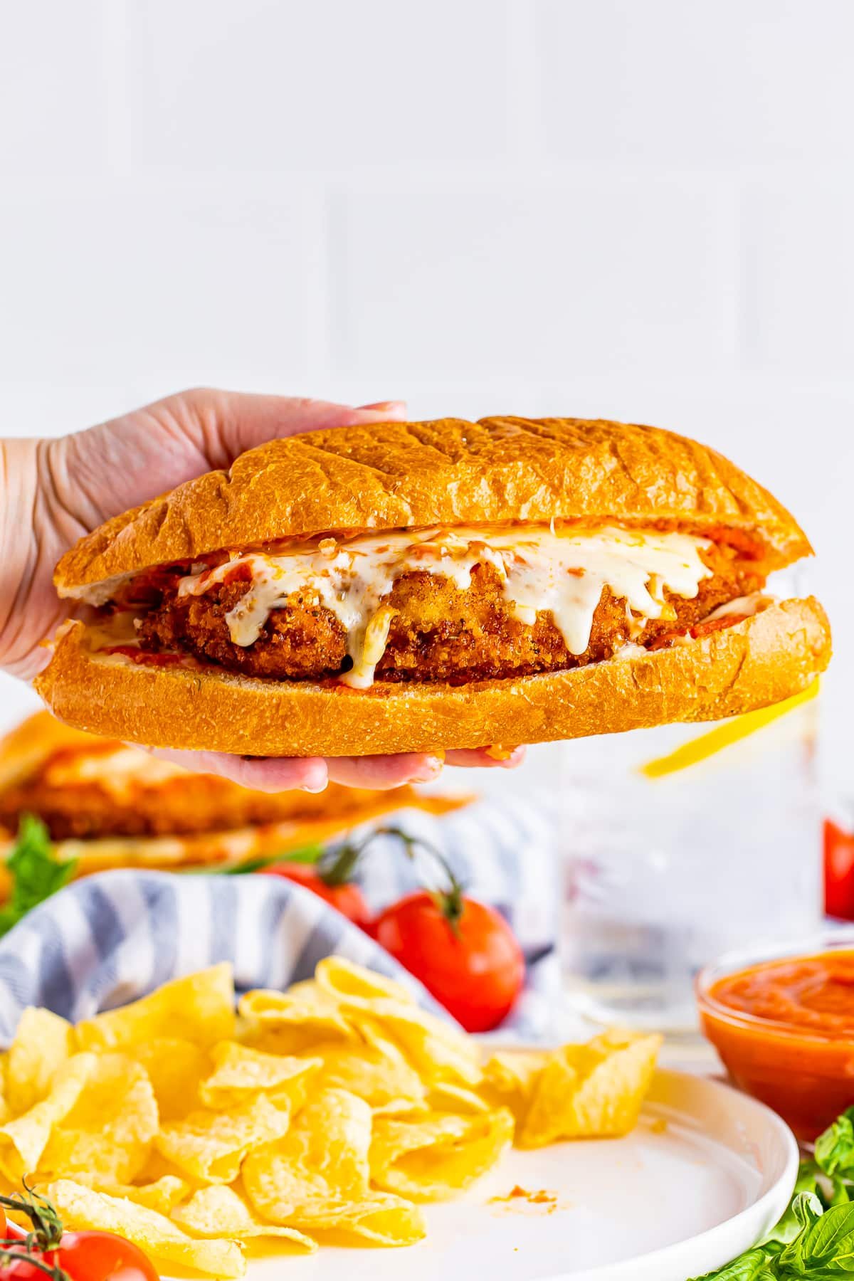 A hand holding a Chicken Parmesan Subs Recipe in mid air with white plate with chips on it, tomatoes, blue striped linen in background