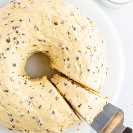 A whole chocolate chip bundt cake with cookie dough frosting on a white cake stand, one slice cut.