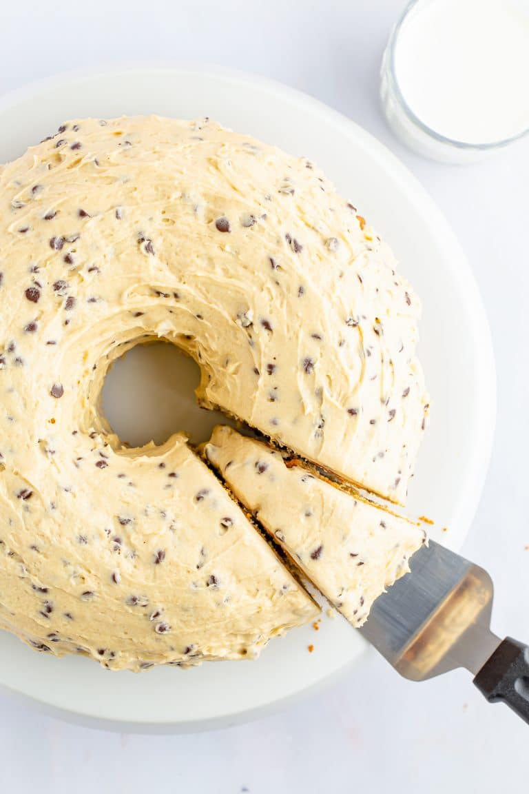 A whole chocolate chip bundt cake with cookie dough frosting on a white cake stand, one slice cut.