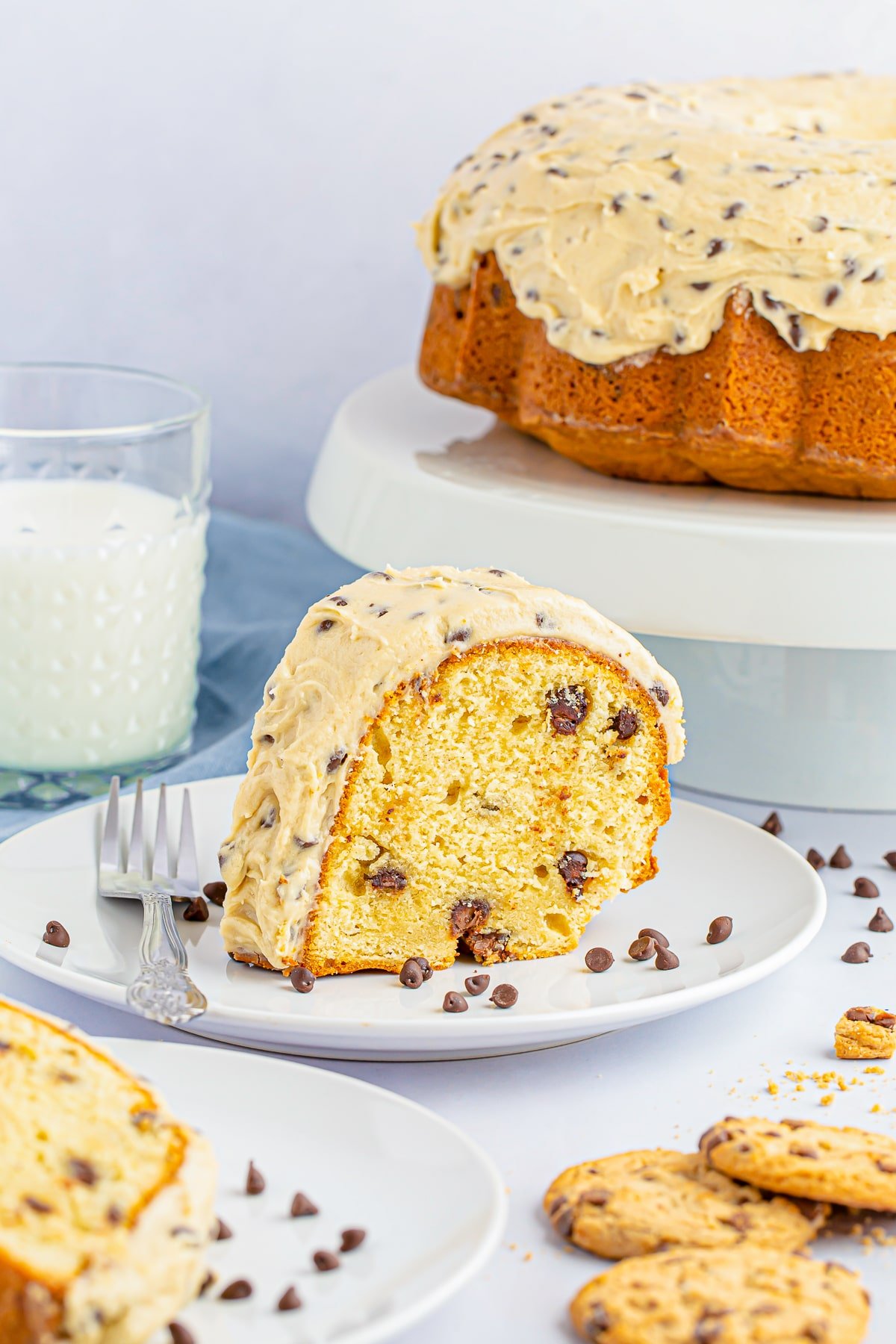 Slice of chocolate chip bundt cake on a white plate with silver fork, light blue linen in the background, glass of milk and bundt cake on cake stand
