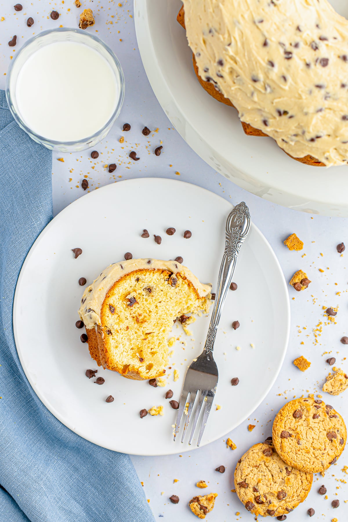 Overhead image of a slice of chocolate chip bundt cake on a white plate, the remainder of the cake on a white cake stand, glass of milk, blue linen and cookie crumbs