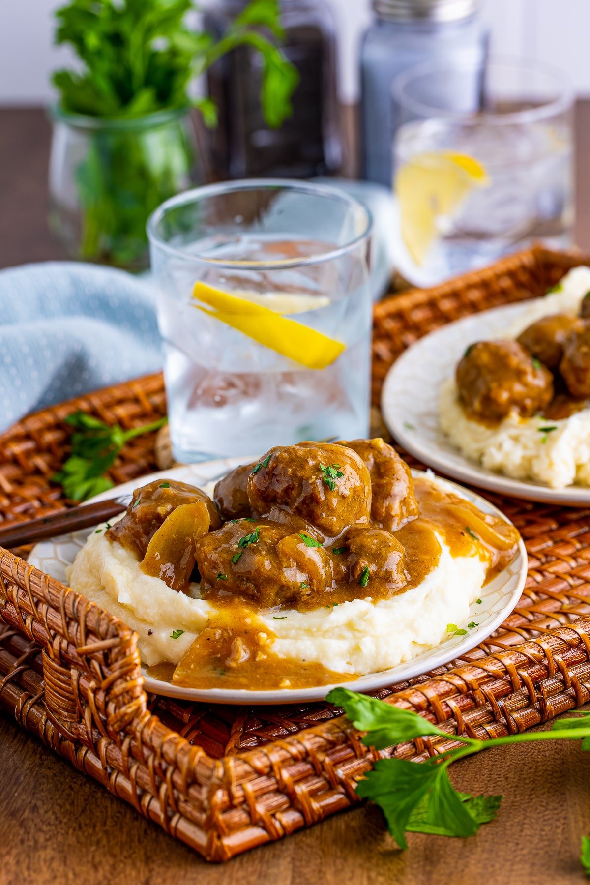 Straight on image showing Meatballs and Gravy served over mashed potatoes on a white plate on a wicker tray. Glass of iced water with lemon in the background with light blue linen, parlsey, and salt and pepper shakers.