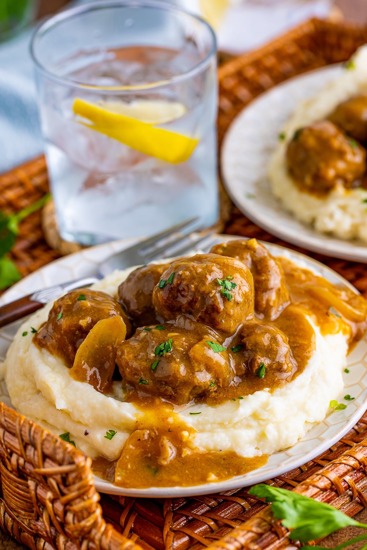 A plate of Meatballs and gravy served over mashed potatoes on a wicker tray, glass of ice water with lemon in the background and light blue linen.