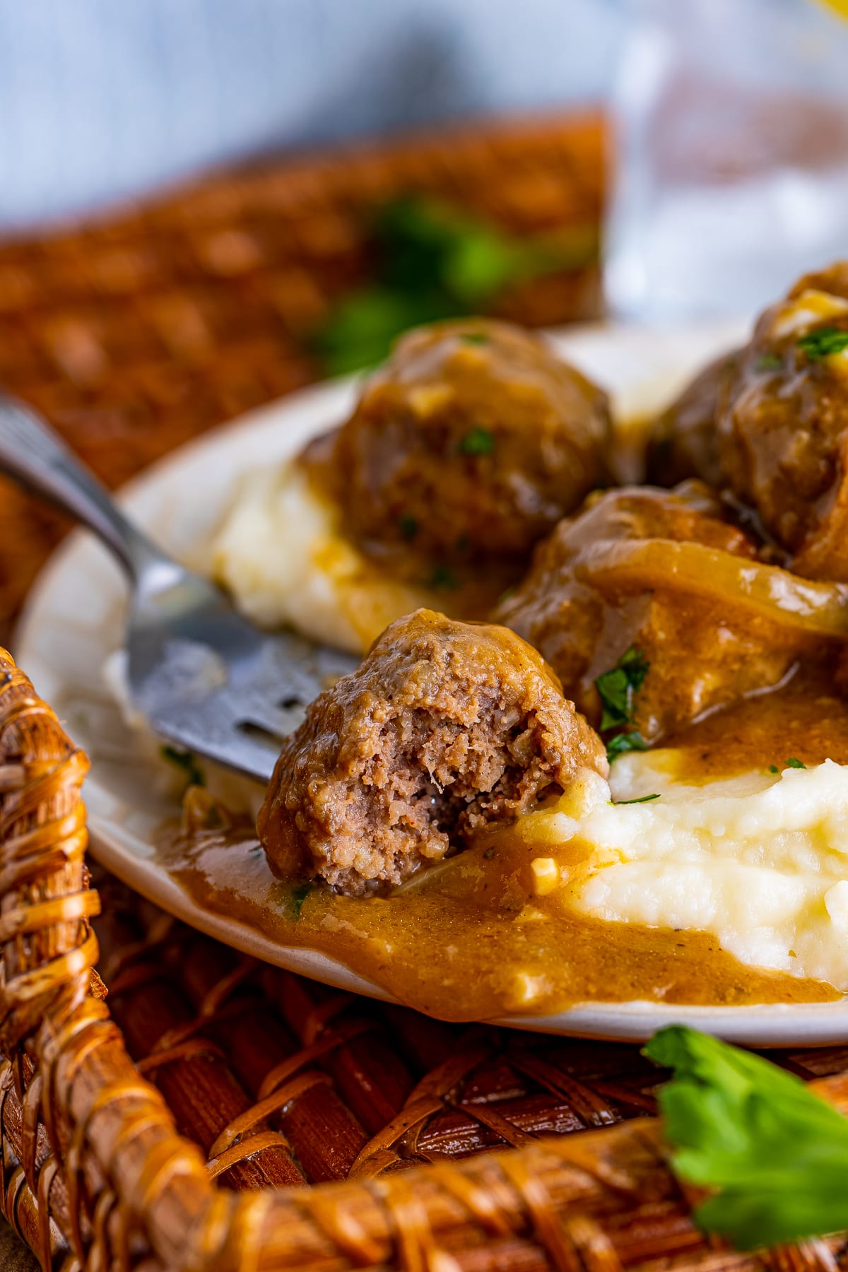 Close up interior shot of a bite of Meatballs and Gravy on a white plate over mashed potatoes. Plate is sitting on a wicker tray with light blue linen and parsley.