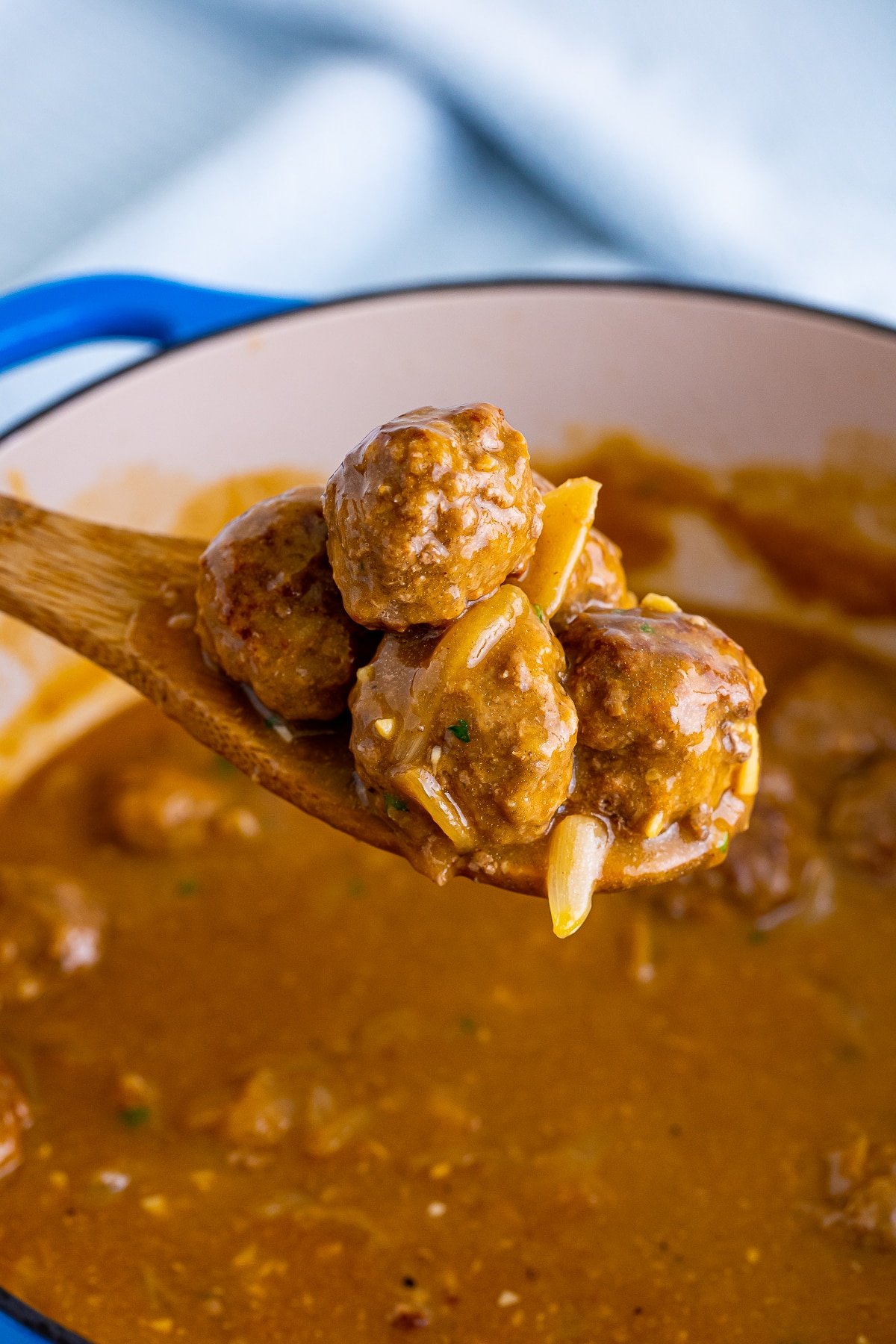 A wooden spoon holding up Meatballs and Gravy in a blue dutch oven with a light blue linen in the background.