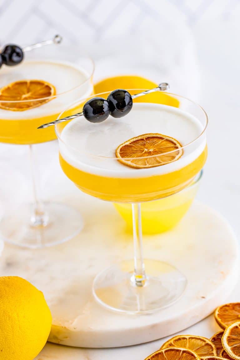 A rum sour in a coupe glass with a silky white egg white foam top, dried lemon wheel, and skewered cherries.