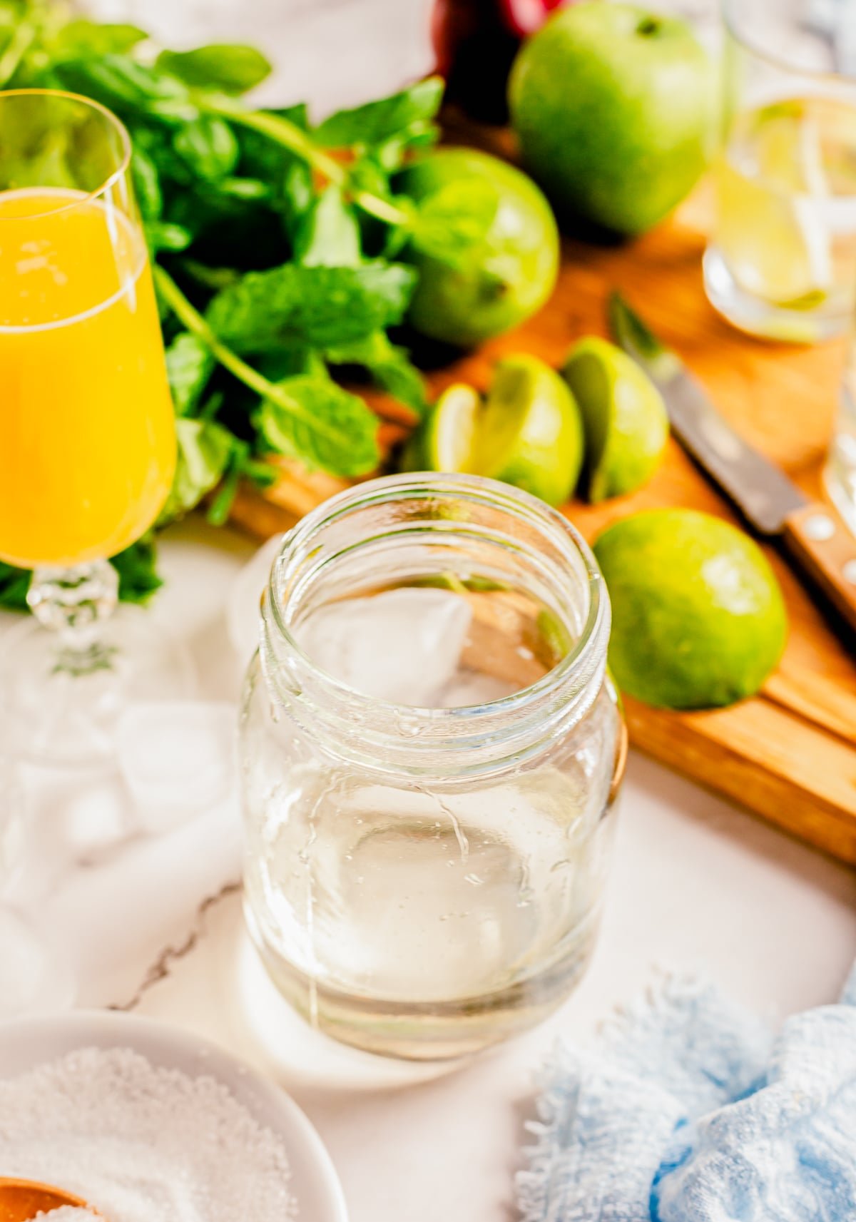 Mason jar with simple syrup for cocktails in it, with a mimosa, limes, mint, wood cutting board and knife in the background
