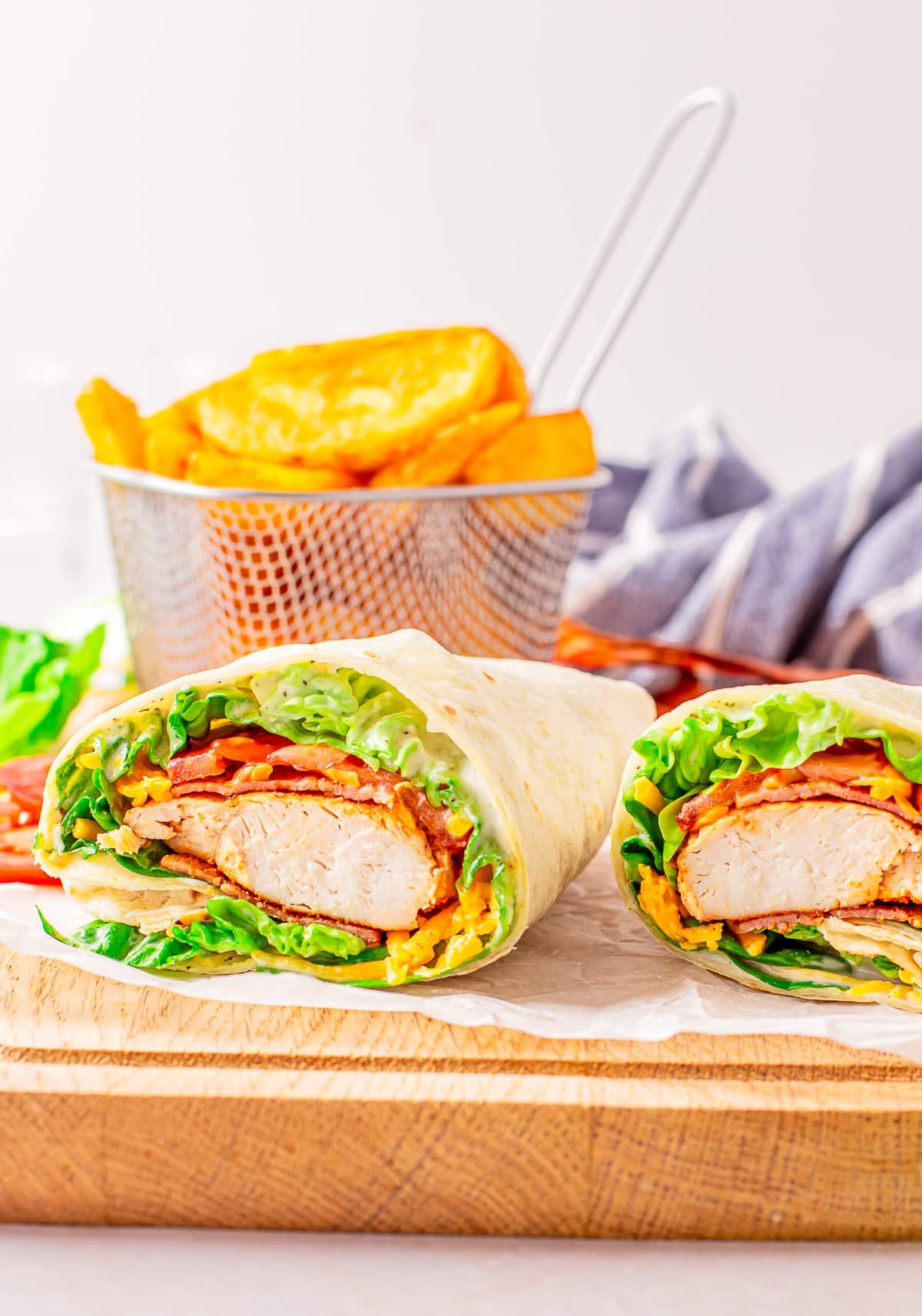 A cut bacon ranch chicken wrap on a piece of white parchment paper sitting on a wooden cutting board. Basket of fries in the background