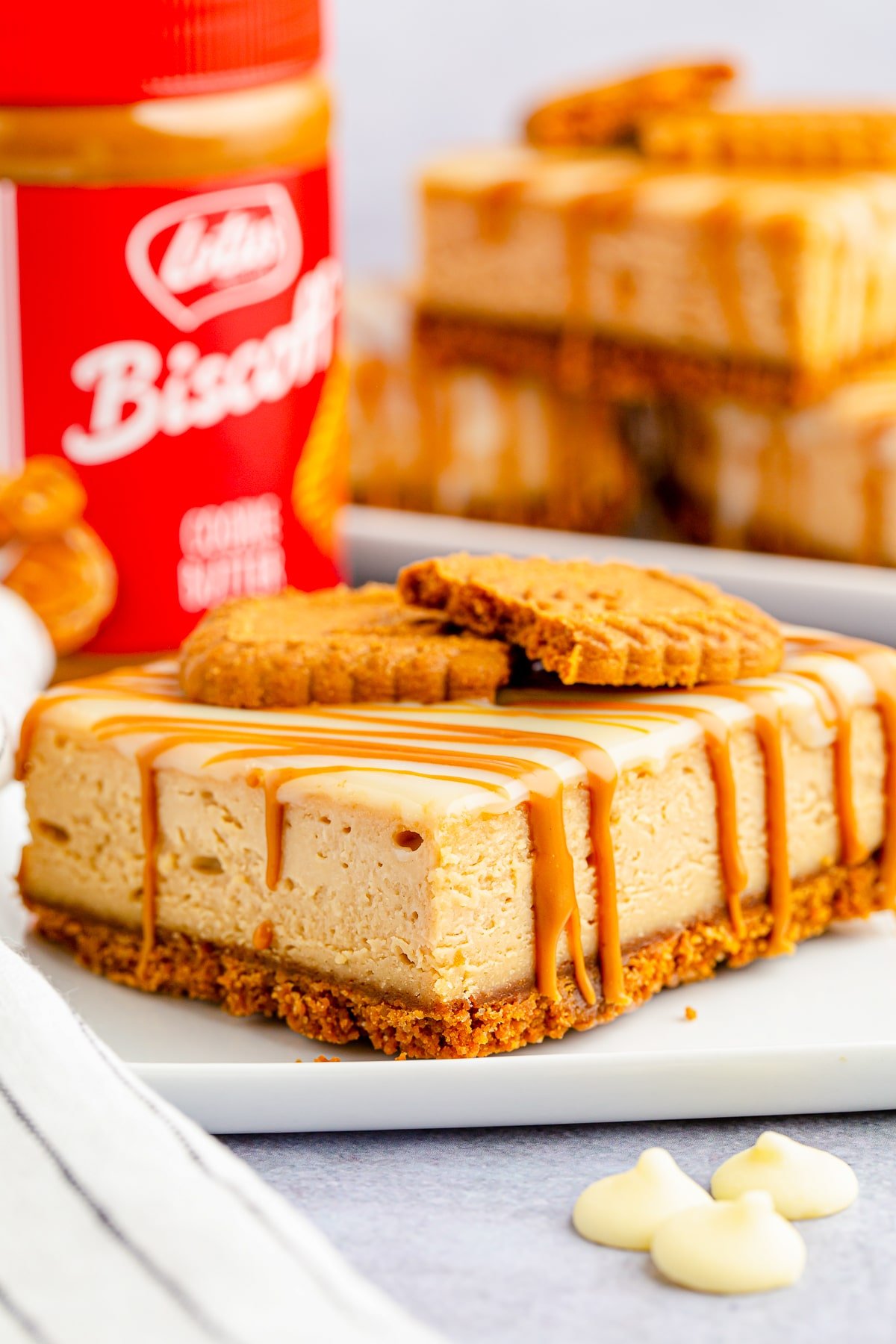A slice of Biscoff Cheesecake Bars on a white plate, jar of cookie butter in the background
