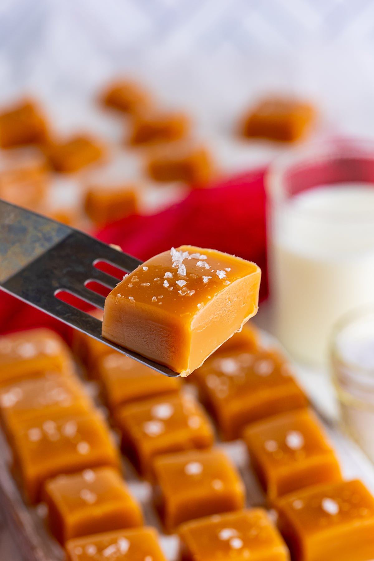 A Salted Caramel Recipe piece on a spatula hovering in air with more pieces in the background, glass of milk, and red linen