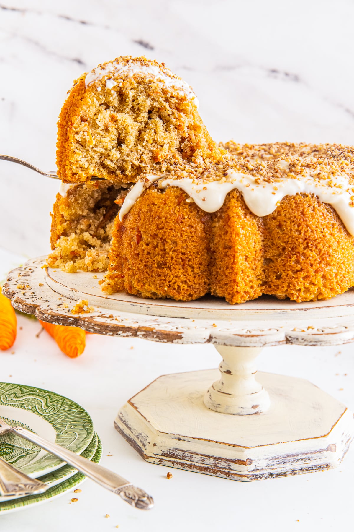 A slice of carrot bundt cake being lifted out by a cake server on a ivory wooden cake stand.