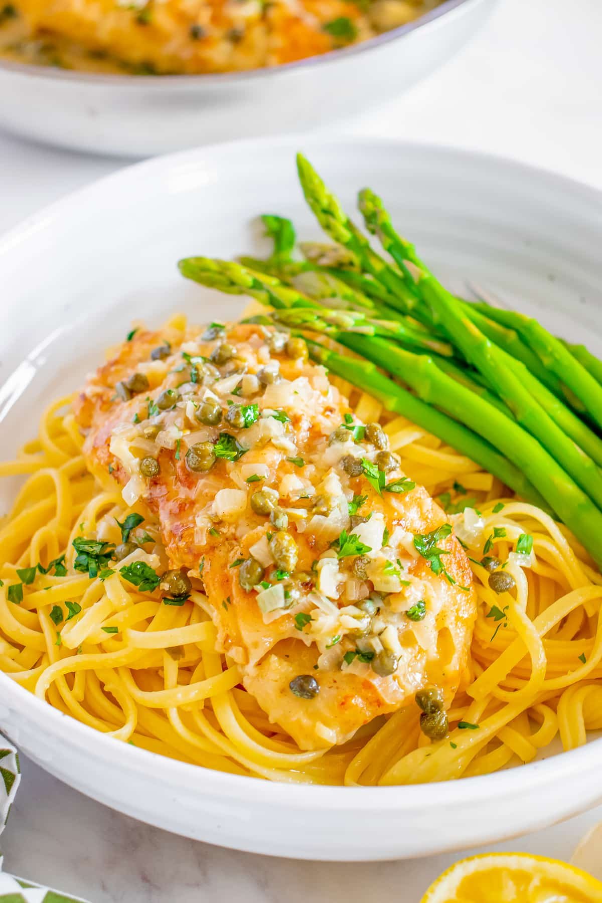 Fish piccata shown served over pasta in a white bowl with asparagus
