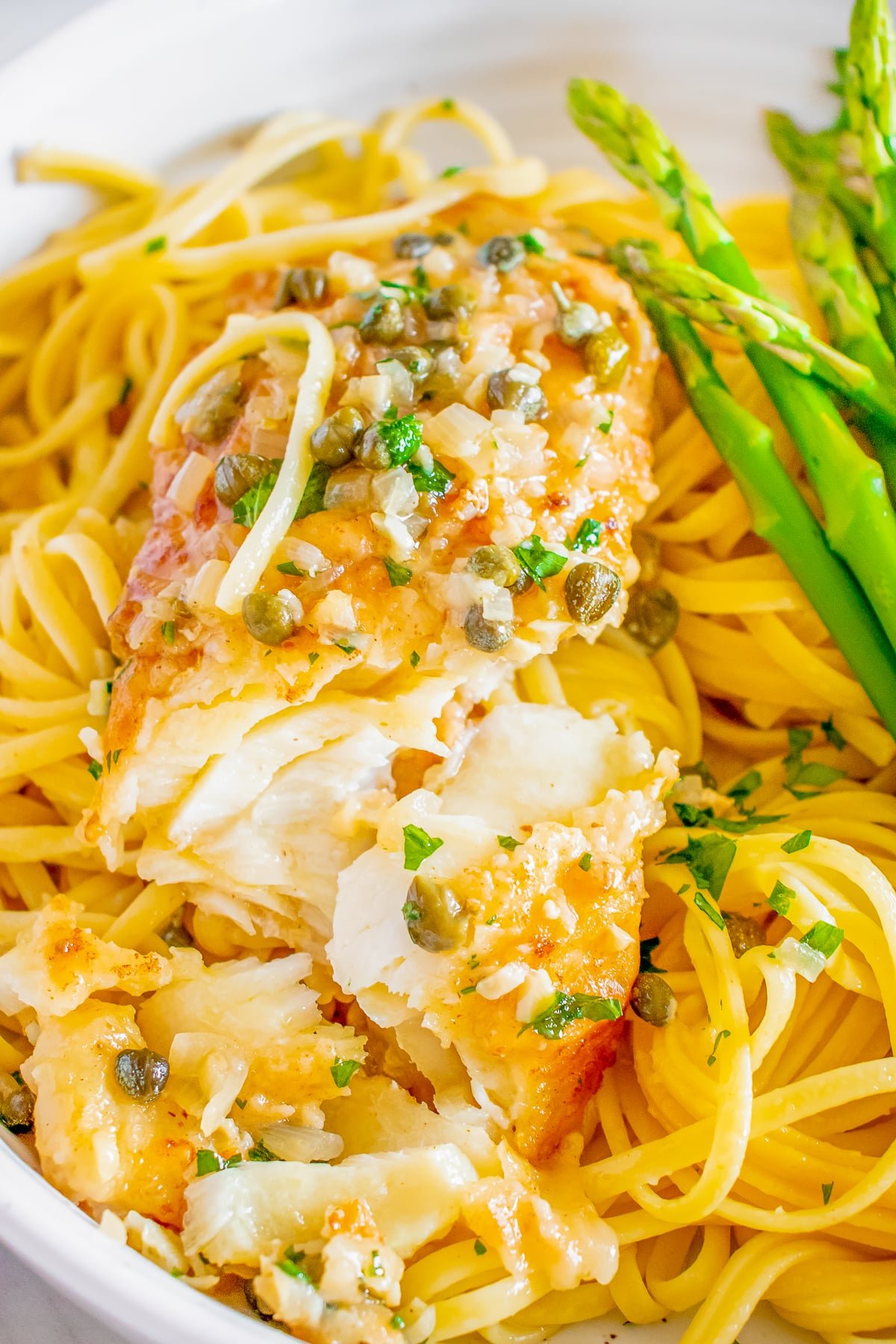 Fish piccata on a bed of pasta, flaked fish showing interior with a side of asparagus