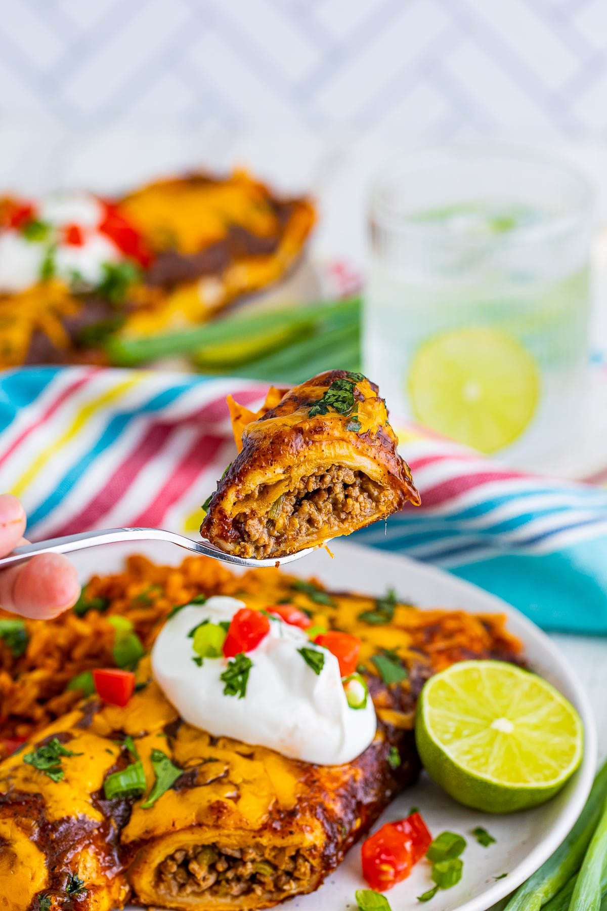 A piece of wet burrito recipe on a silver fork in air, colorful linen, iced water and plate with more burritos in the background