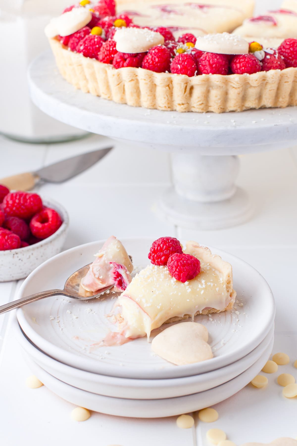 A slice of Raspberry Tart on a stack of white plates with with a piece taken out with a fork. White cake stand with the tart on it in the back ground, white while countertop, bowl of raspberries and cake knife in background.