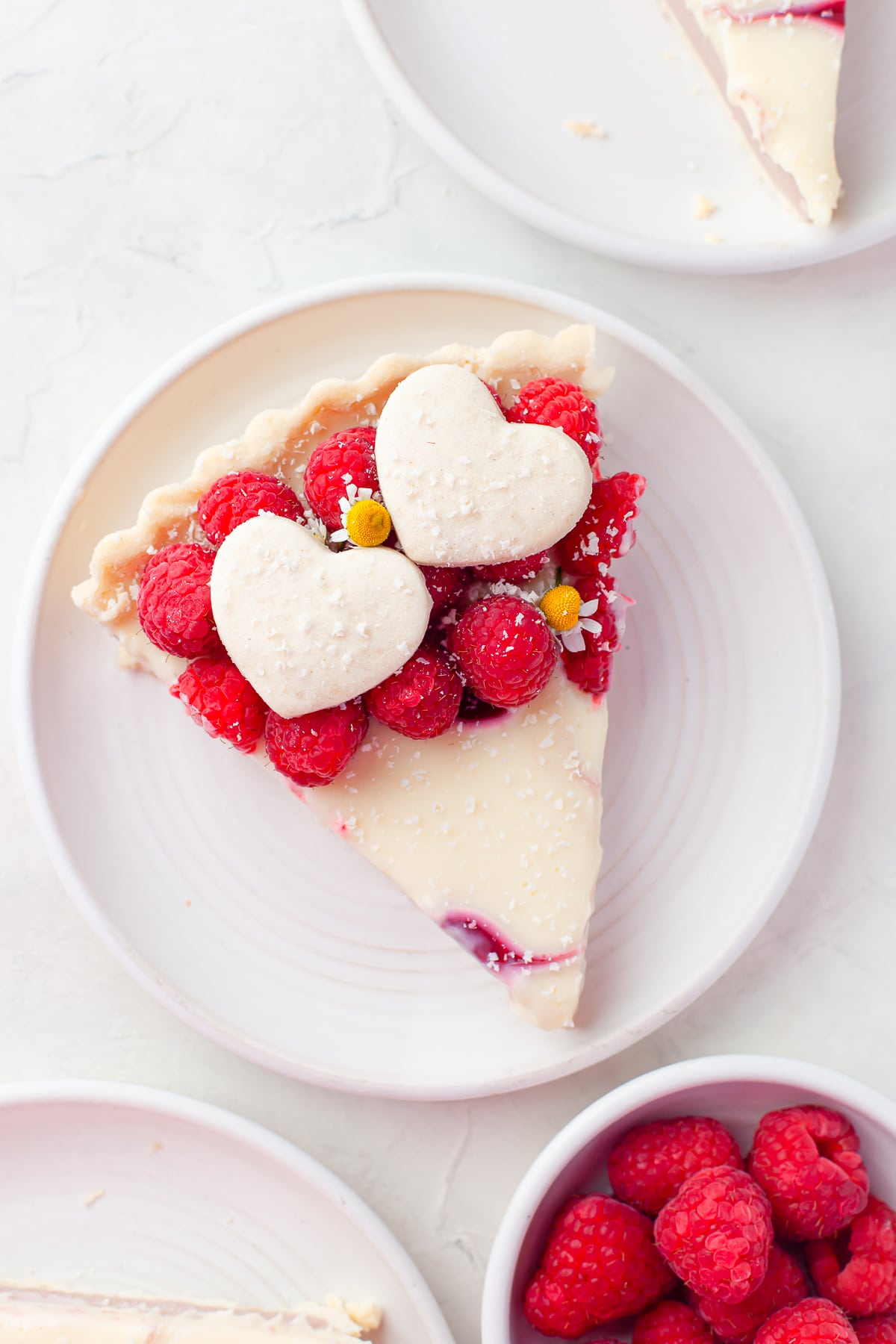 Overhead shot of a slice of Raspberry Tart on a white plate on white countertop with bowl of raspberries