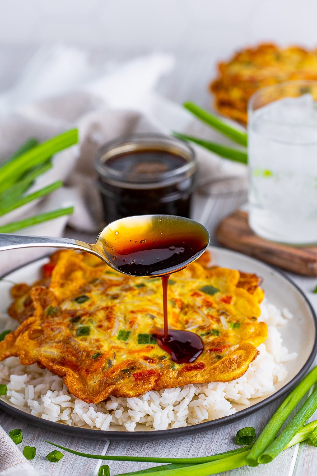 Vegetable Egg Foo Young Recipe on a plate with the gravy being drizzled on top with a silver spoon. Glass of ice water, scallions, and jar of sauce in the background