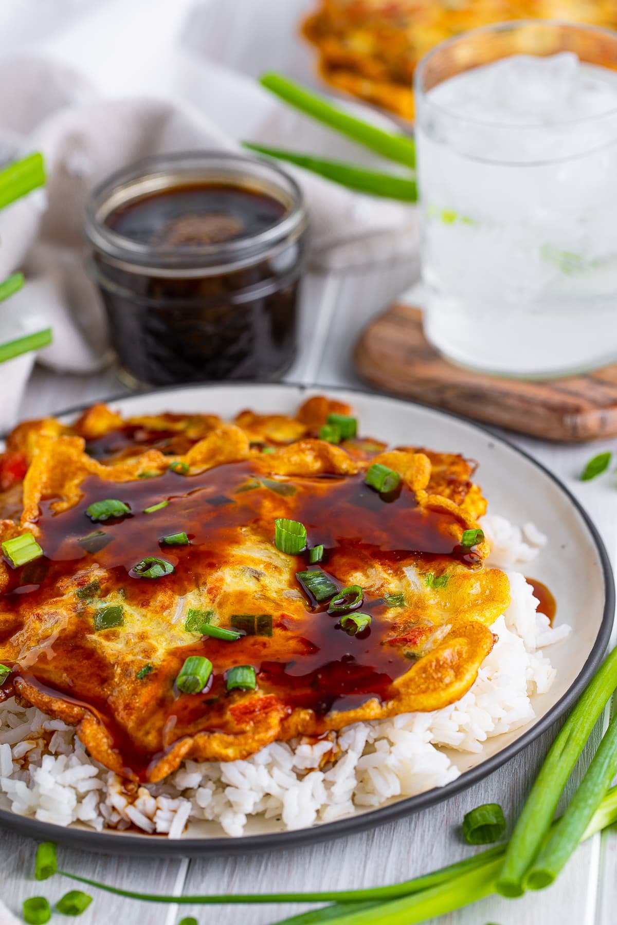 Vegetable Egg Foo Young Recipe on a white wooden table top with glass of ice water, scallions, and jar of sauce in the background.