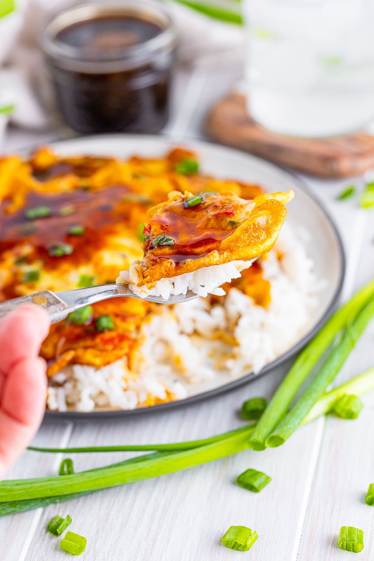 A fork holding up a bite of Vegetable Egg Foo Young Recipe with the plate blurred in the background with jar of sauce, ice water, and scallions