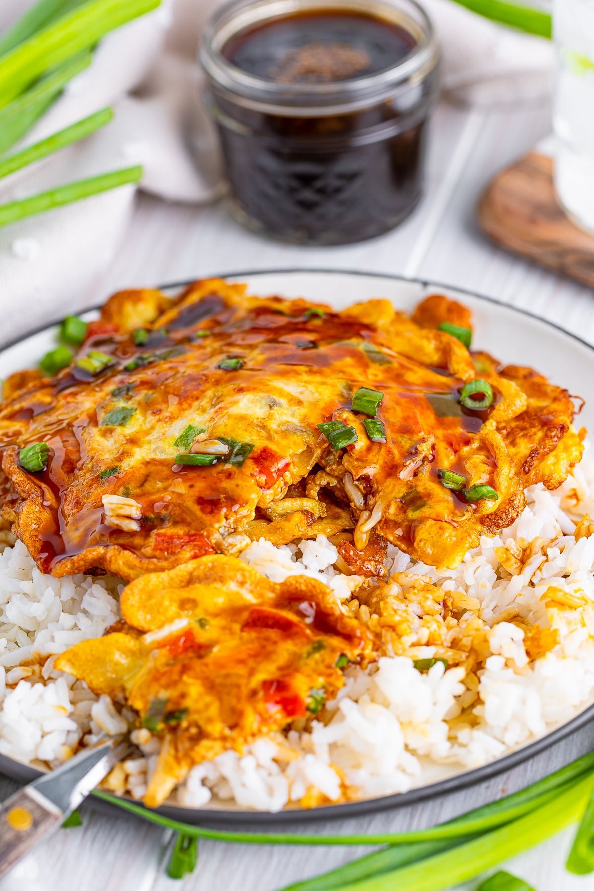 A piece of Vegetable Egg Foo Young recipe taken out of one pancake on a bed of white rice, on a white wooden tabletop, jar of sauce and scallions in the background