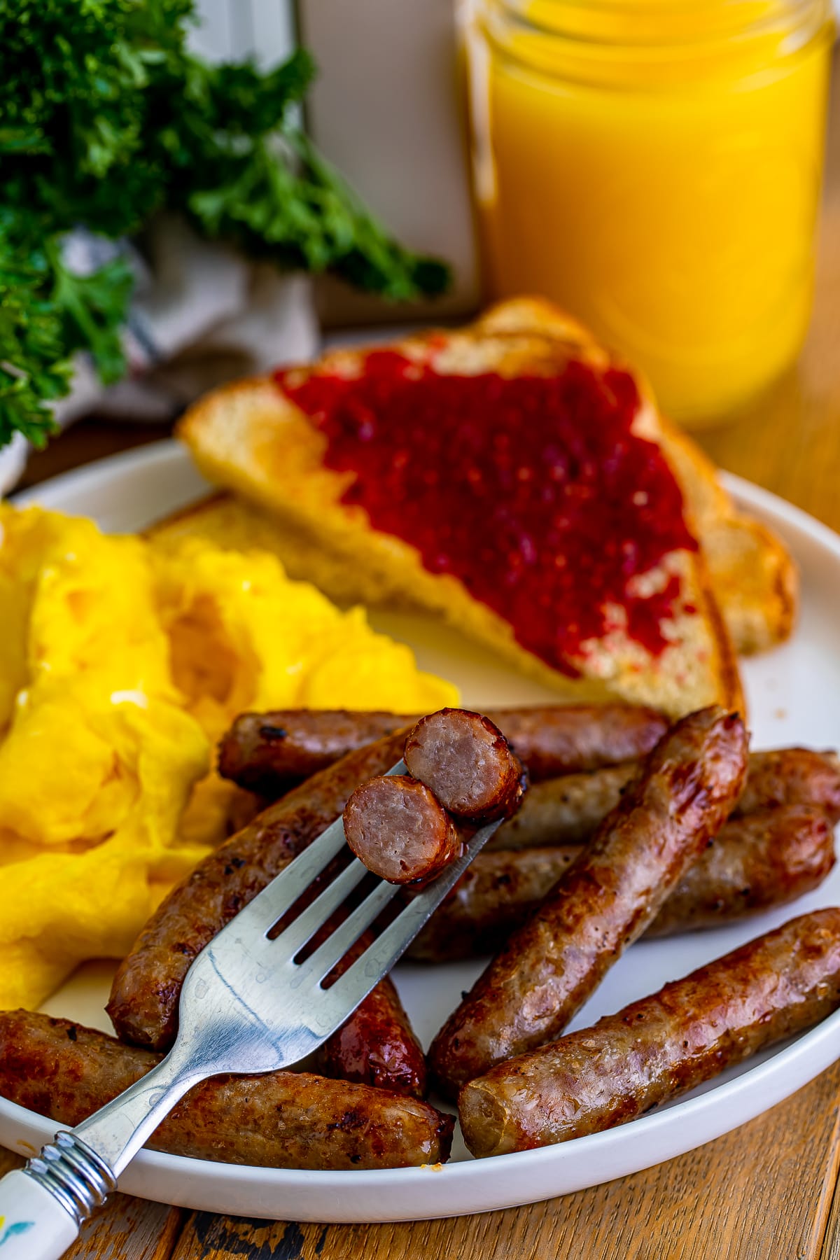 Air fryer breakfast sausage links on a white plate and on a silver fork with toast and jam in the background with scrambled eggs