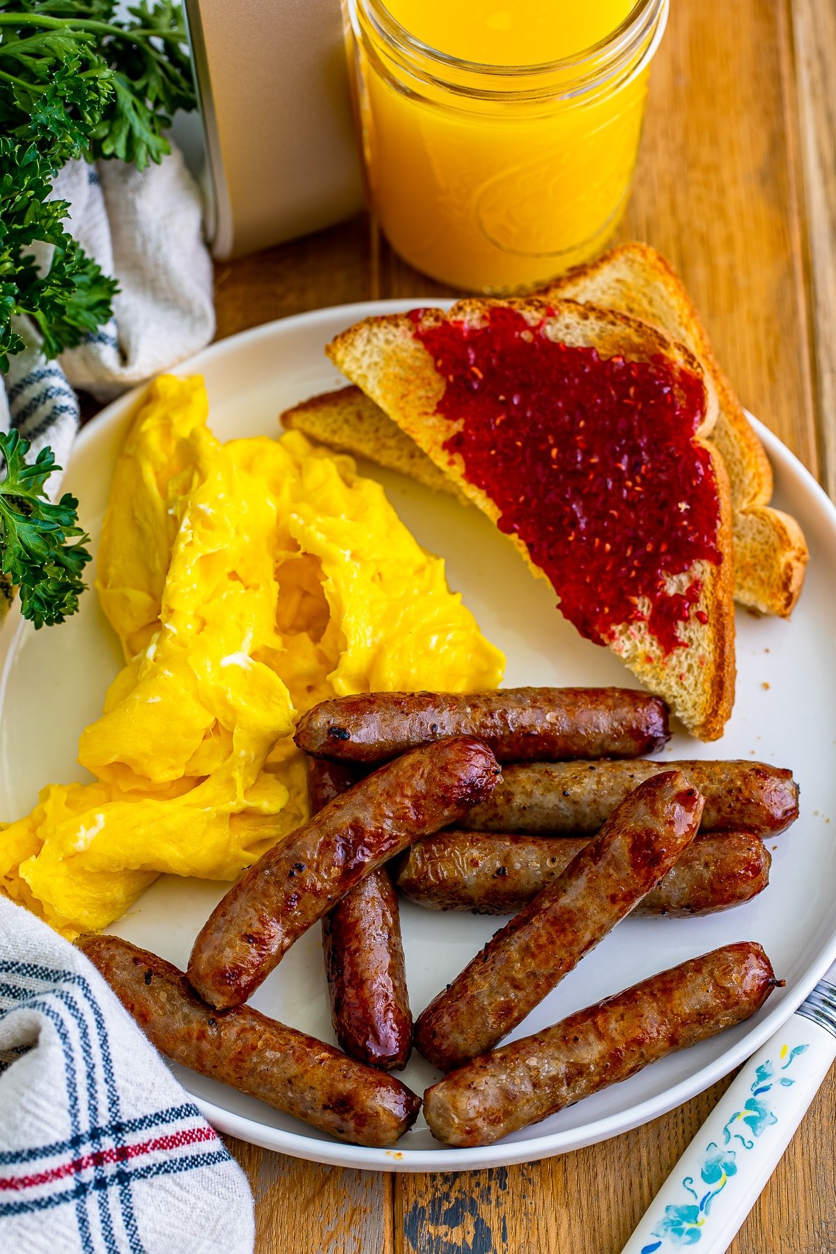 Overhead image of air fryer breakfast sausage links on a white plate with toast and jam, scrambled eggs, on a wooden table top with orange juice