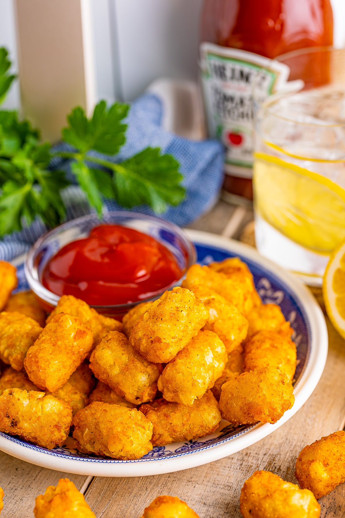 The best crispy air fryer tater tots on a blue plate on a wooden table top with glass of iced water with lemon, blue check linen, parsley, and ketchup