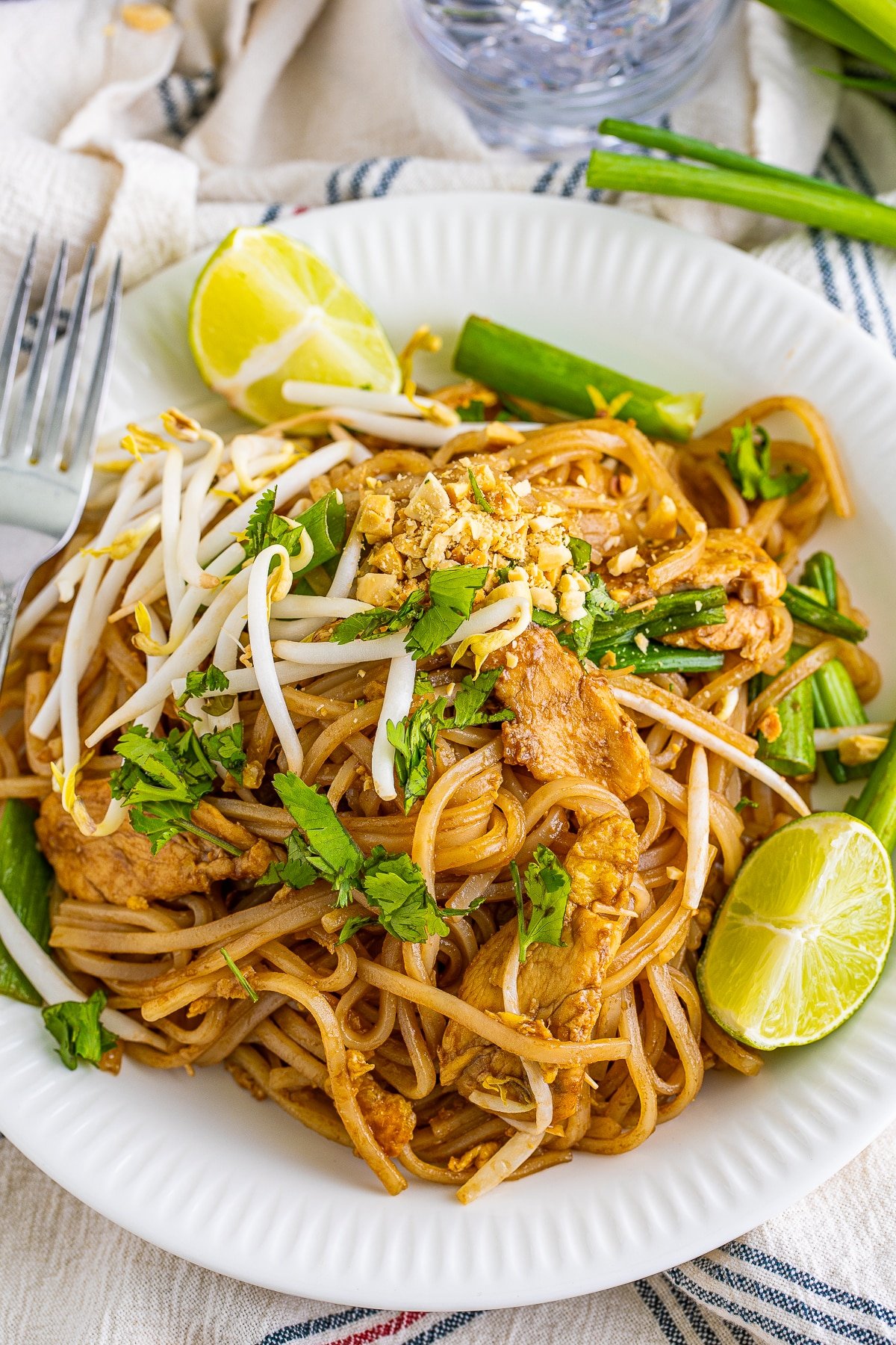 Overhead image showing spicy pad thai recipe on a white plate with silver fork, limes, and scallions