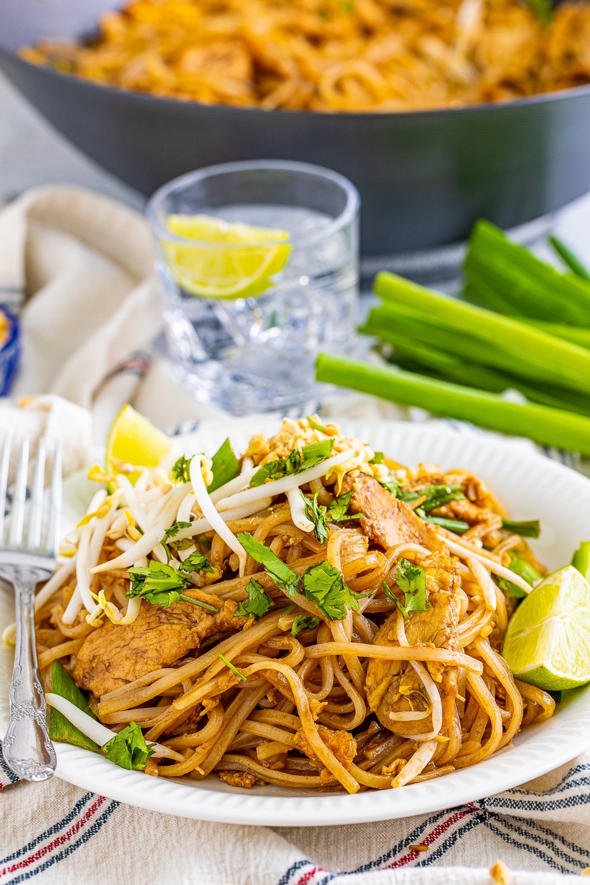 A further away image of spicy pad Thai recipe on a white plate with silver fork, green onion, iced water, and wok in the background