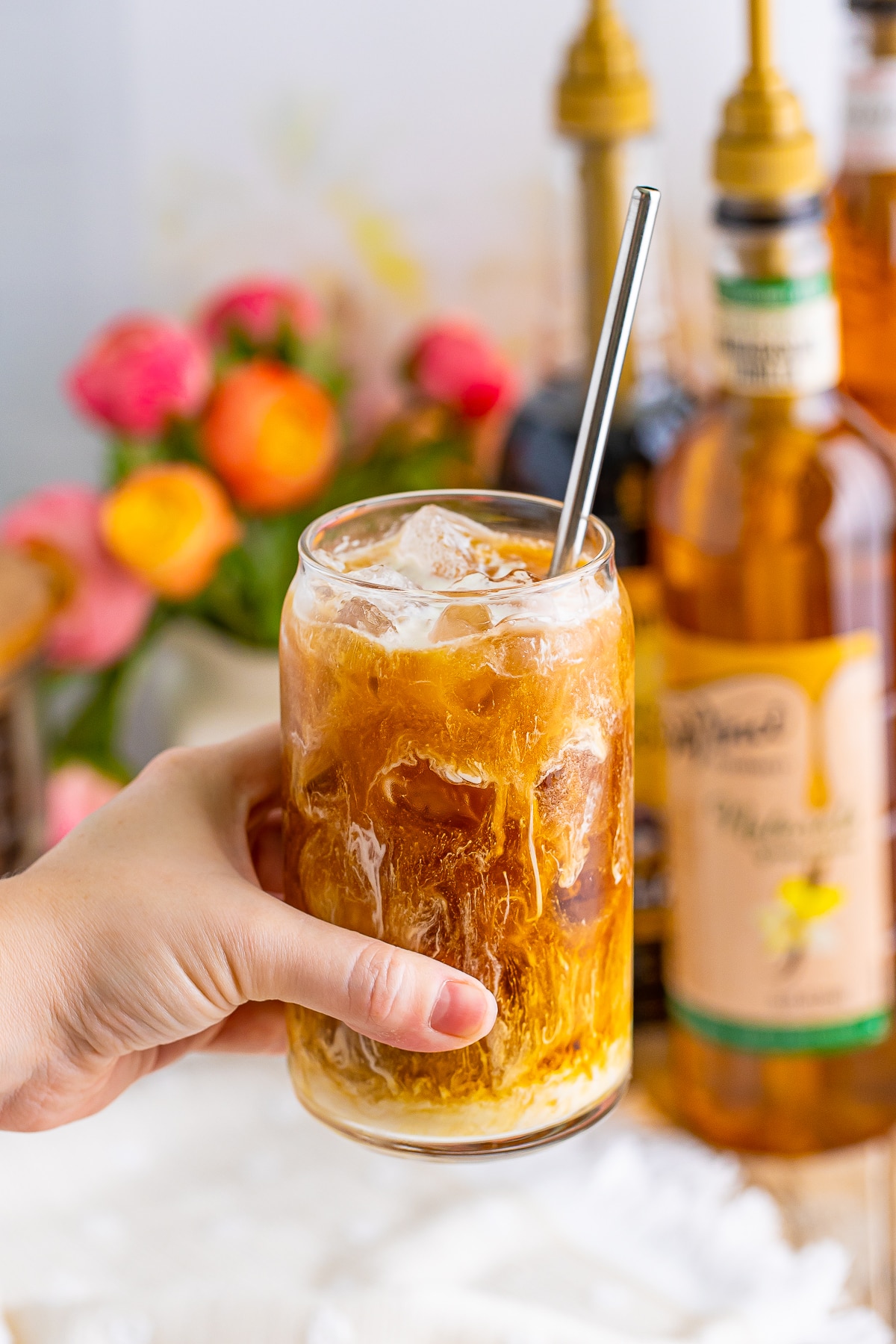 a hand holding an iced coffee in a glass with cream and a metal straw, coffee syrups in the background and flowers