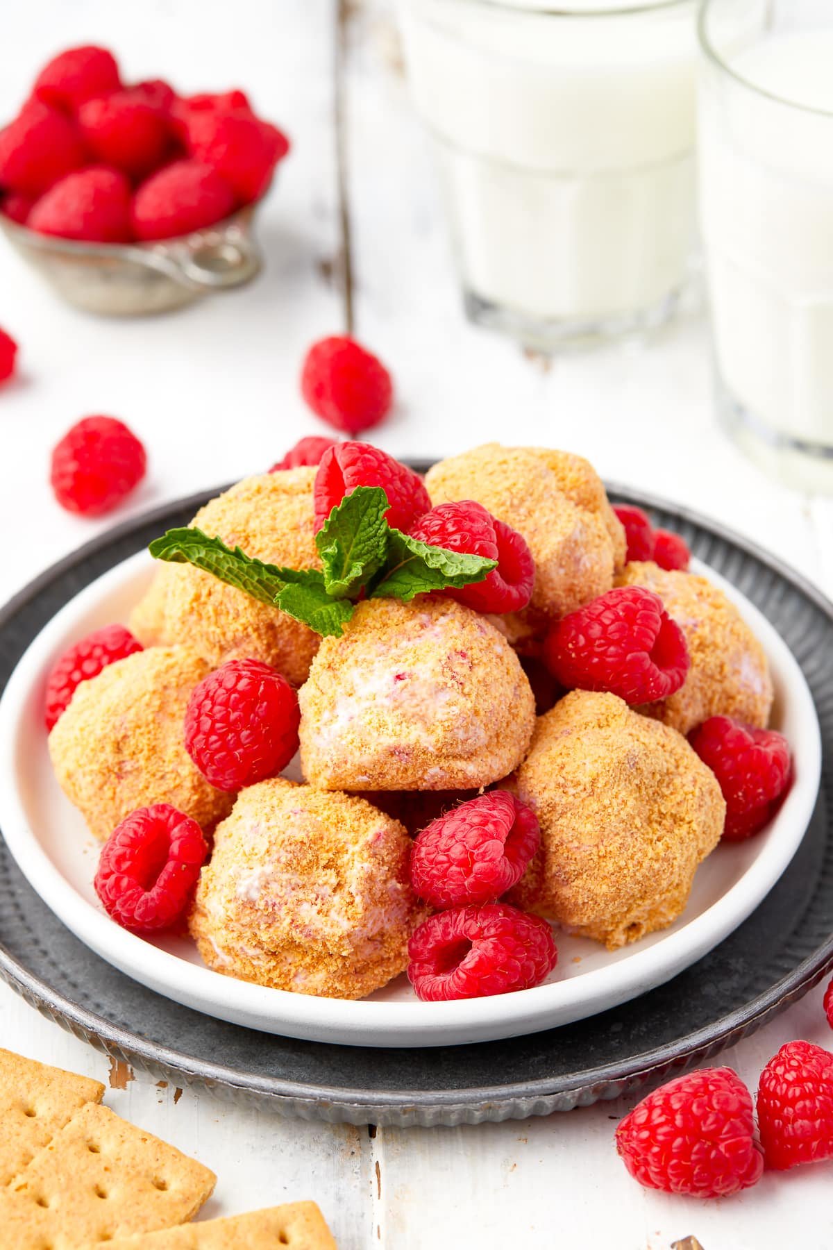 No bake cheesecake bites on a white plate sitting on a metal serving platter with fresh raspberries and min