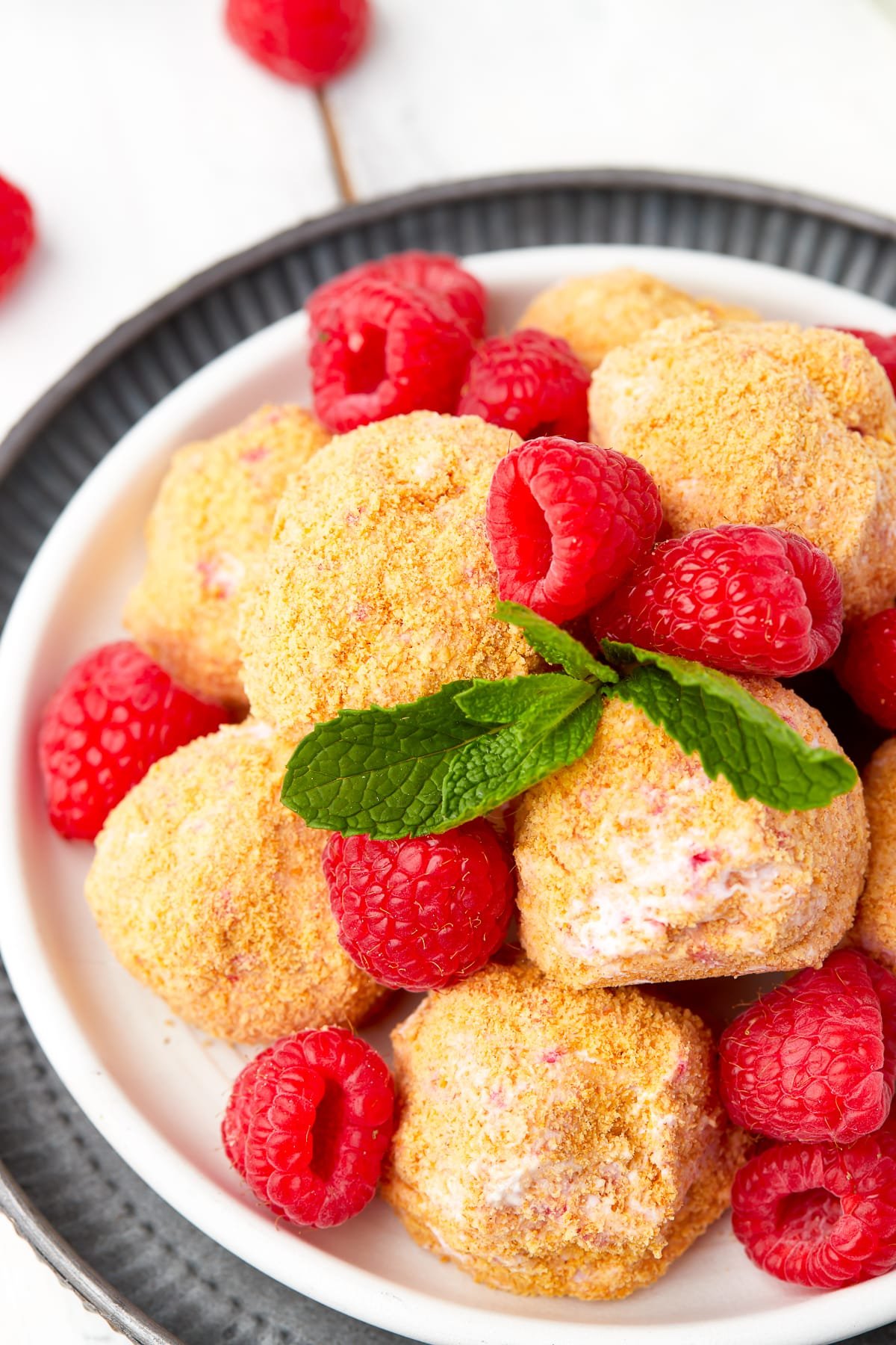 No bake cheesecake bites on a white plate sitting on a metal serving platter with fresh raspberries and mint